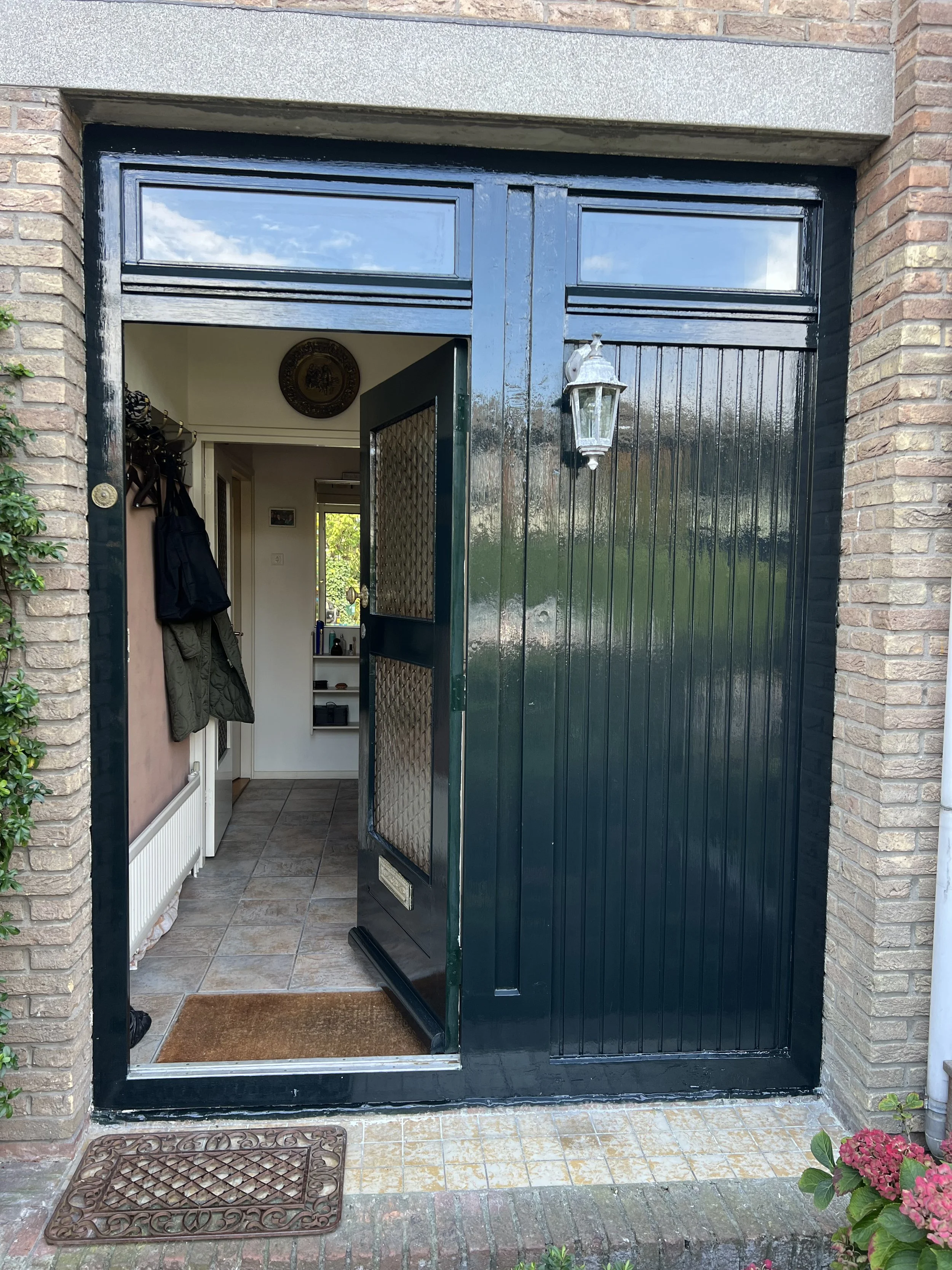 De ingang van een huis met een open deur en een zwarte houten deur aan de rechterkant. Binnen is de gang zichtbaar met een kapstok met jassen en tassen. Buiten liggen twee deurmatten, één voor de deur en één in de gang. Aan de rechterkant buiten hangt een muur lampje.