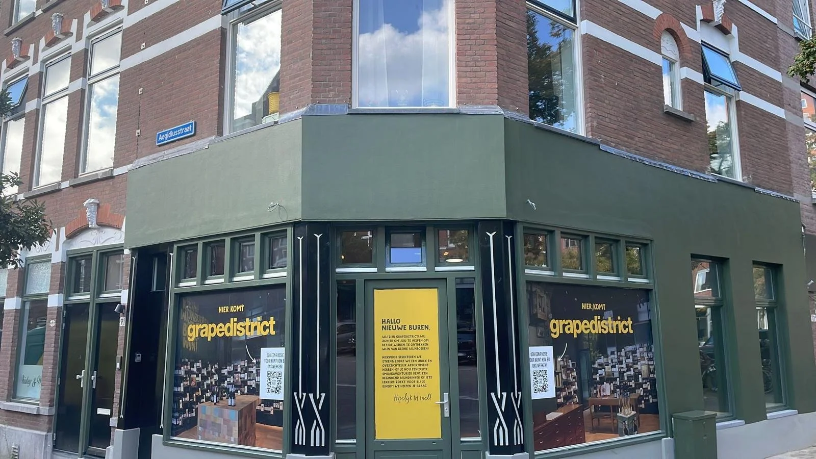 Een hoekpand met een groene gevel en grote ramen, met posters in de ramen voor Grapedistrict, een winkel die zich richt op wijn, met een gele deur en reclame. De straatnaam Aegidiusstraat is zichtbaar.