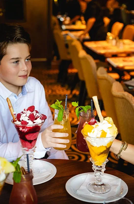Jeune garçon assis à une table dans un restaurant élégant, tenant un cocktail avec des fruits et des feuilles de menthe, entouré de plusieurs autres desserts et boissons colorés.