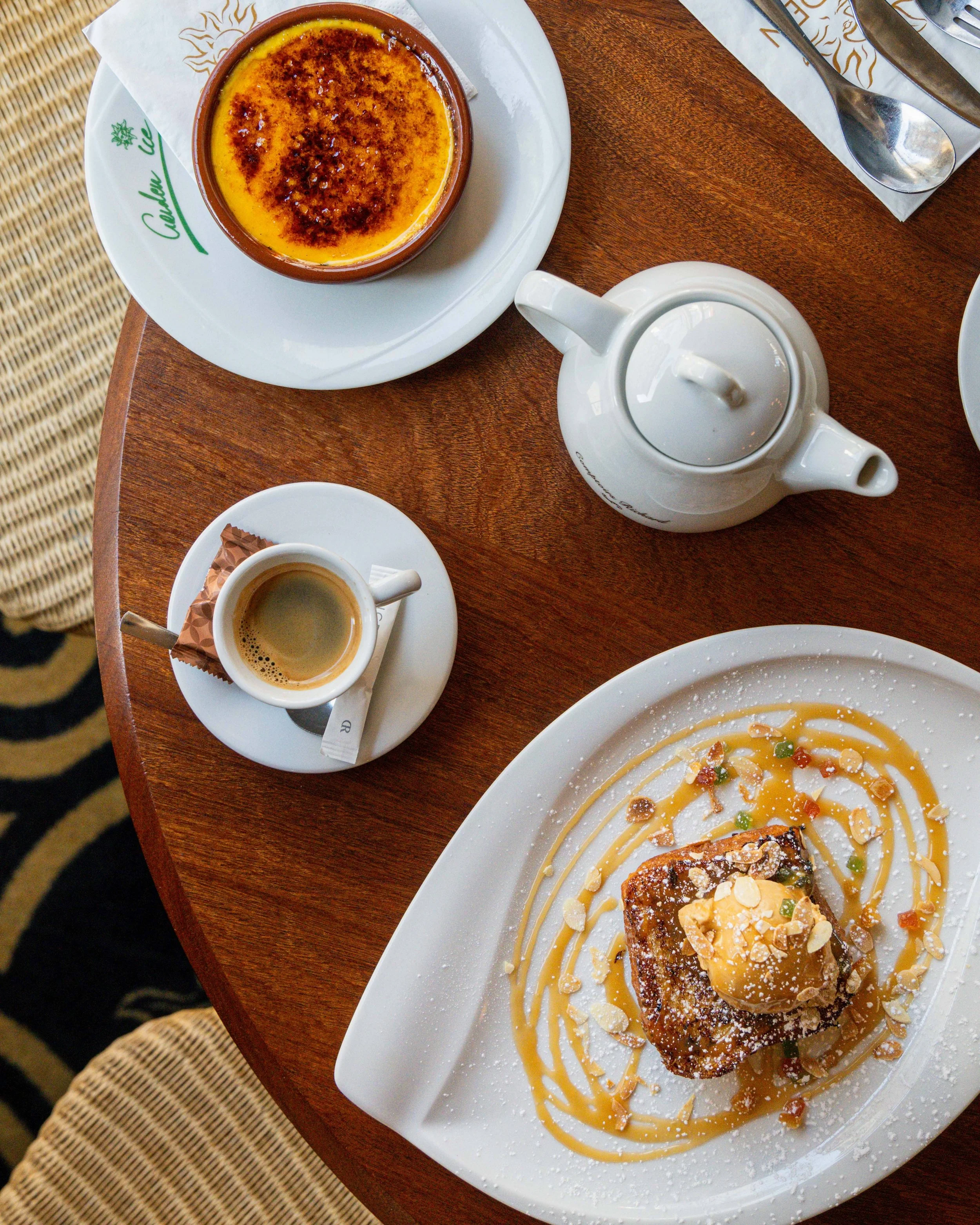 Un dessert crème brûlée dans un ramequin, un petit espresso dans une tasse, une théière blanche, et un plat avec un gâteau garni de caramel, de crème, de noix, et de sucre glace sur une table en bois au Garden Ice Café