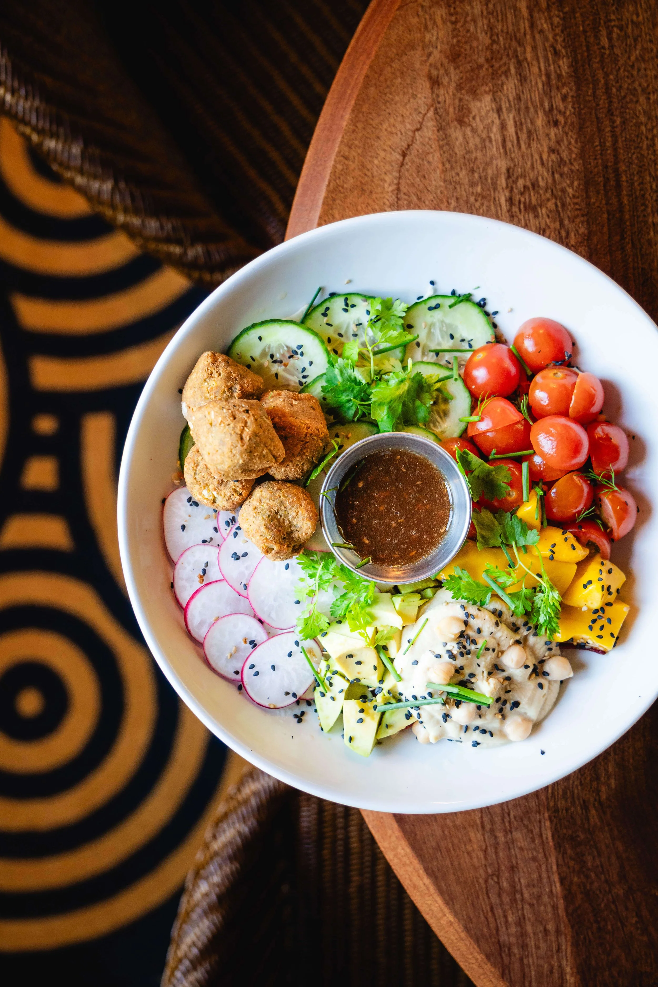 Plat de salade avec concombres, radis, tomates cerises, mangue, avocat, yaourt avec graines de sésame, nuggets, et une sauce au centre, présenté dans une assiette blanche sur une table en bois.