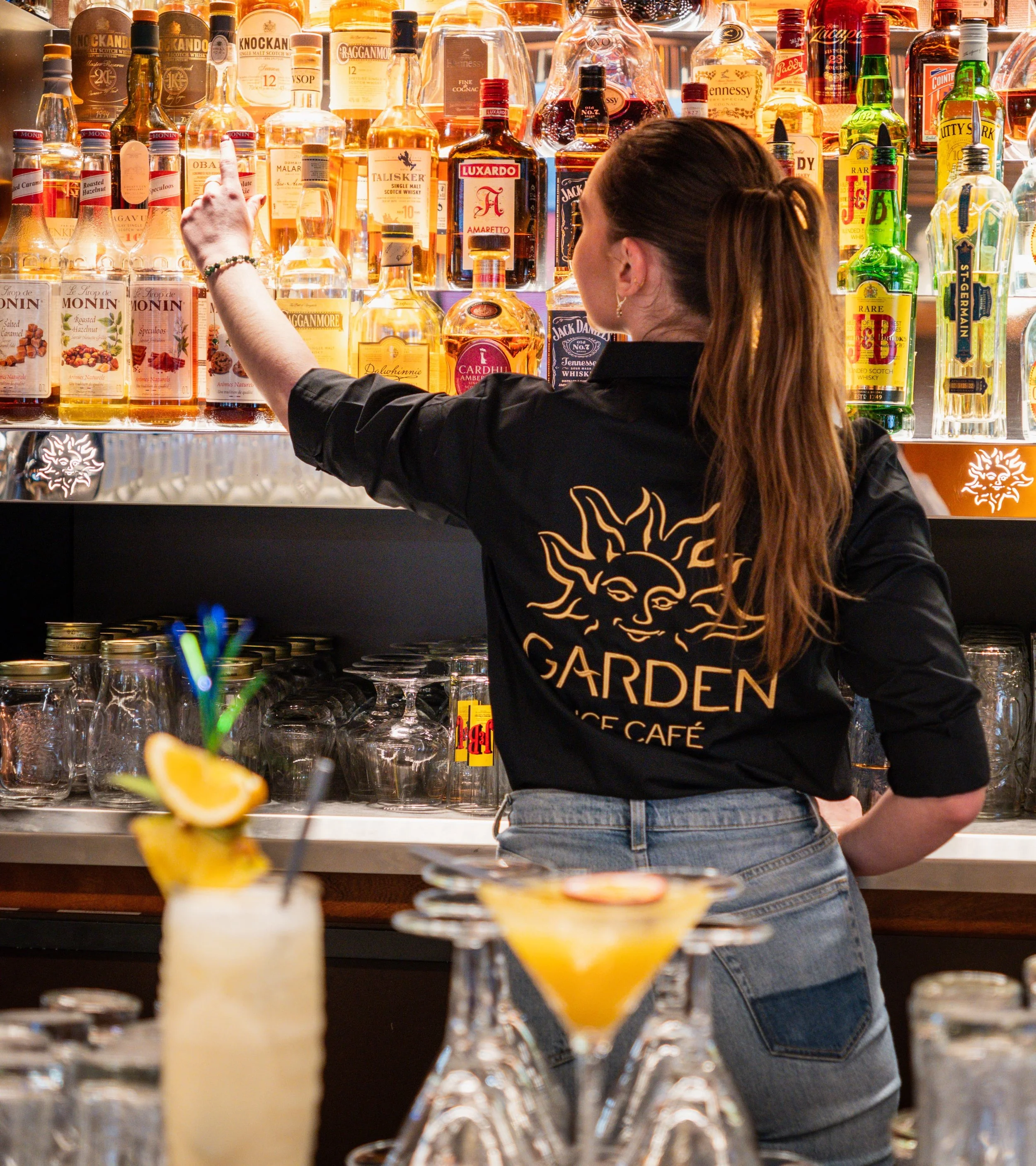 Une barmanne avec une chemise noire portant le logo "Garden Ice" derrière un bar