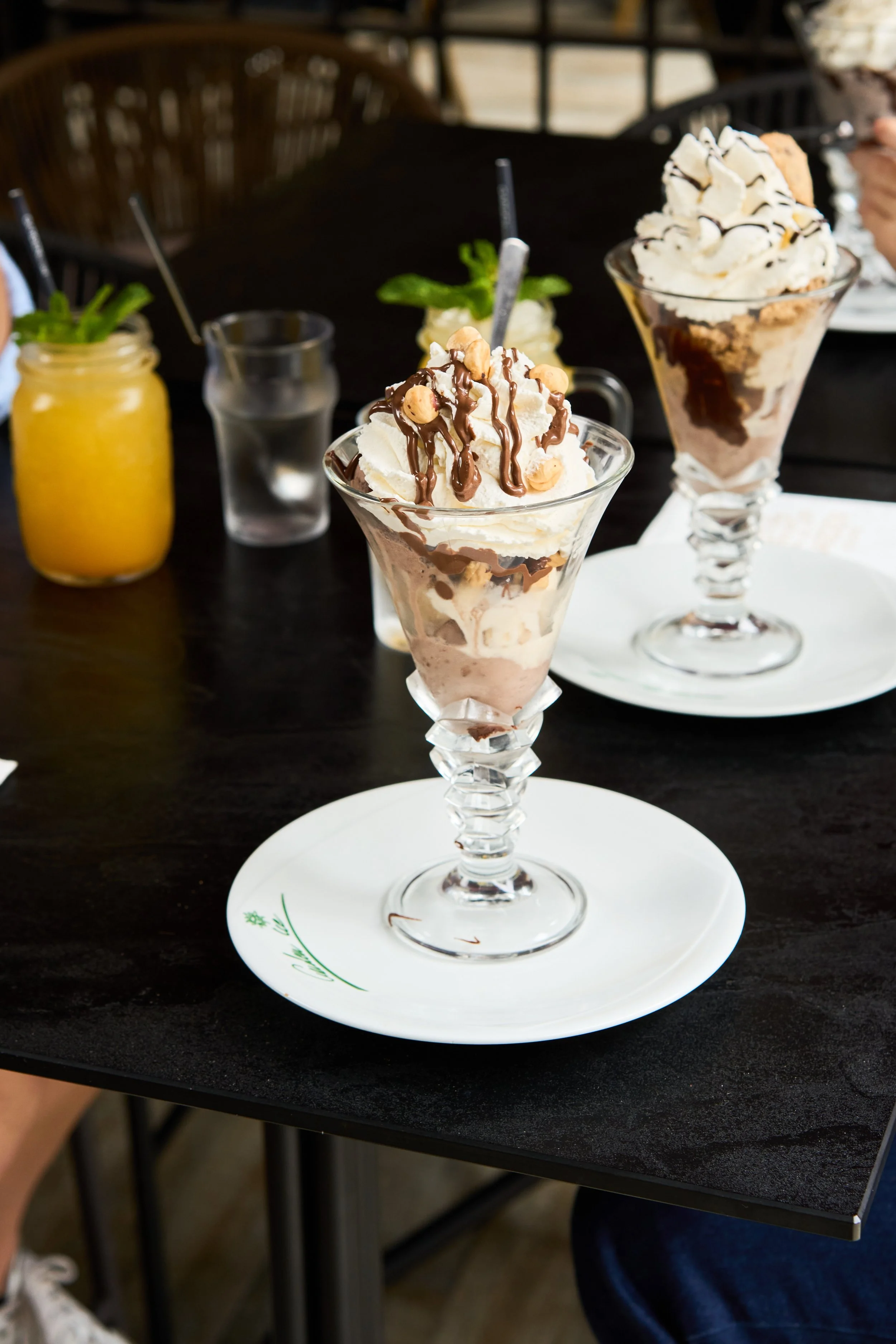 Jeune garçon assis à une table dans un restaurant élégant, tenant un cocktail avec des fruits et des feuilles de menthe, entouré de plusieurs autres desserts et boissons colorés.