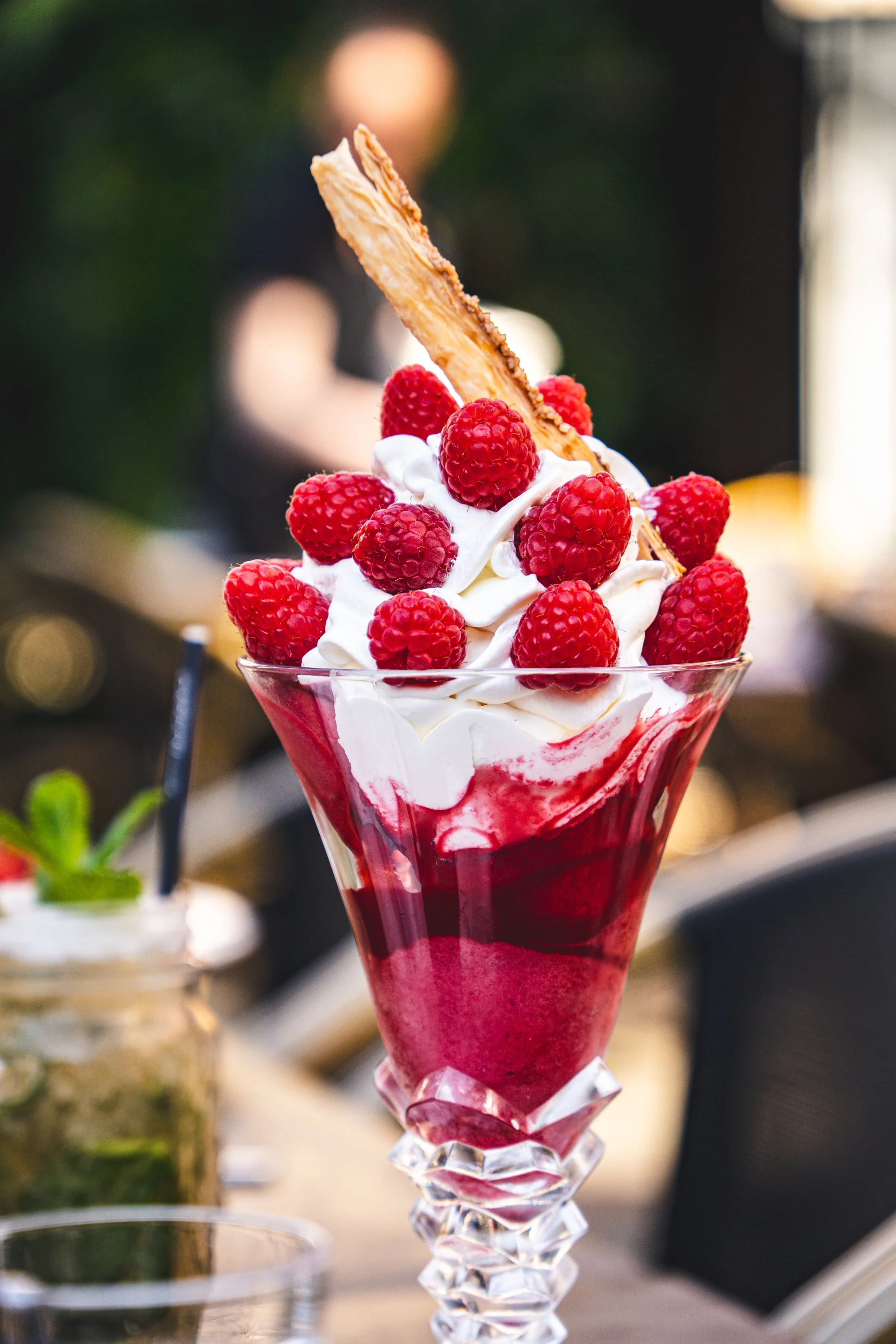 Jeune garçon assis à une table dans un restaurant élégant, tenant un cocktail avec des fruits et des feuilles de menthe, entouré de plusieurs autres desserts et boissons colorés.
