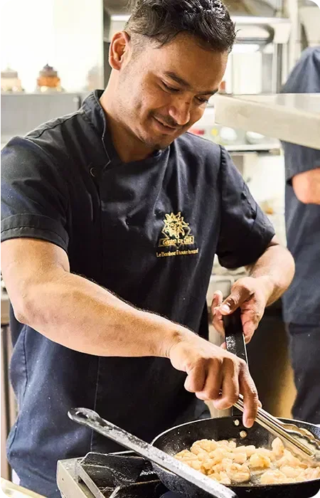 Un chef en cuisine en train de faire sauter des crevettes dans une poêle, portant un uniforme noir avec un logo Garden Ice café