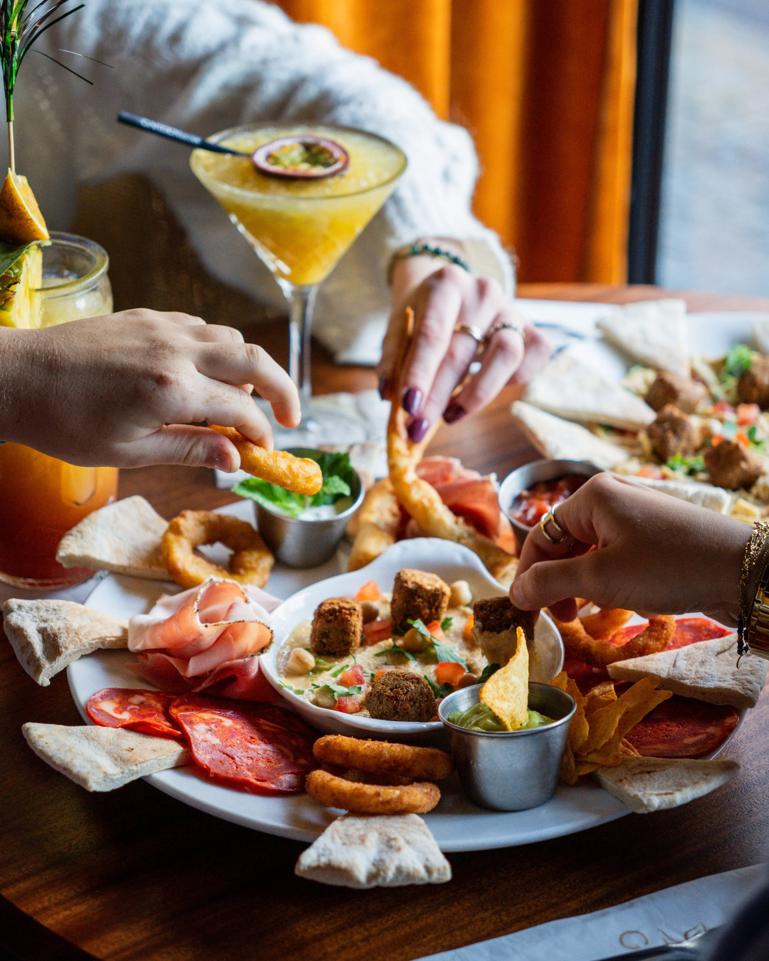 Gros plan d'un plateau de hors-d'œuvre variés, avec des mains en train de prendre des croustilles et des morceaux de pain pita, boissons colorées en arrière-plan, dans une ambiance de restaurant.