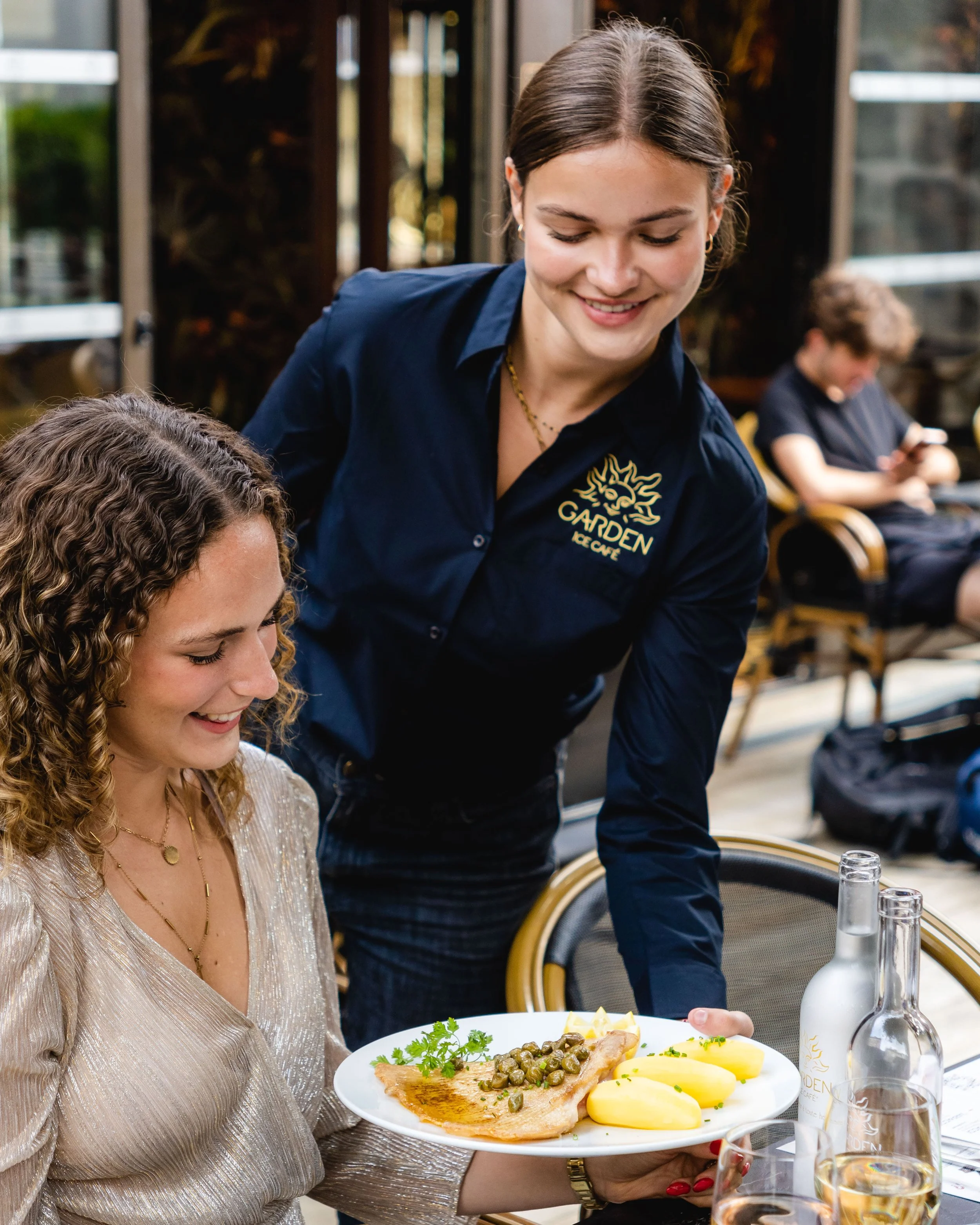 Une femme sert un plat à une femme assise lors d'un repas dans un restaurant, avec d'autres personnes en arrière-plan au Garden Ice Café