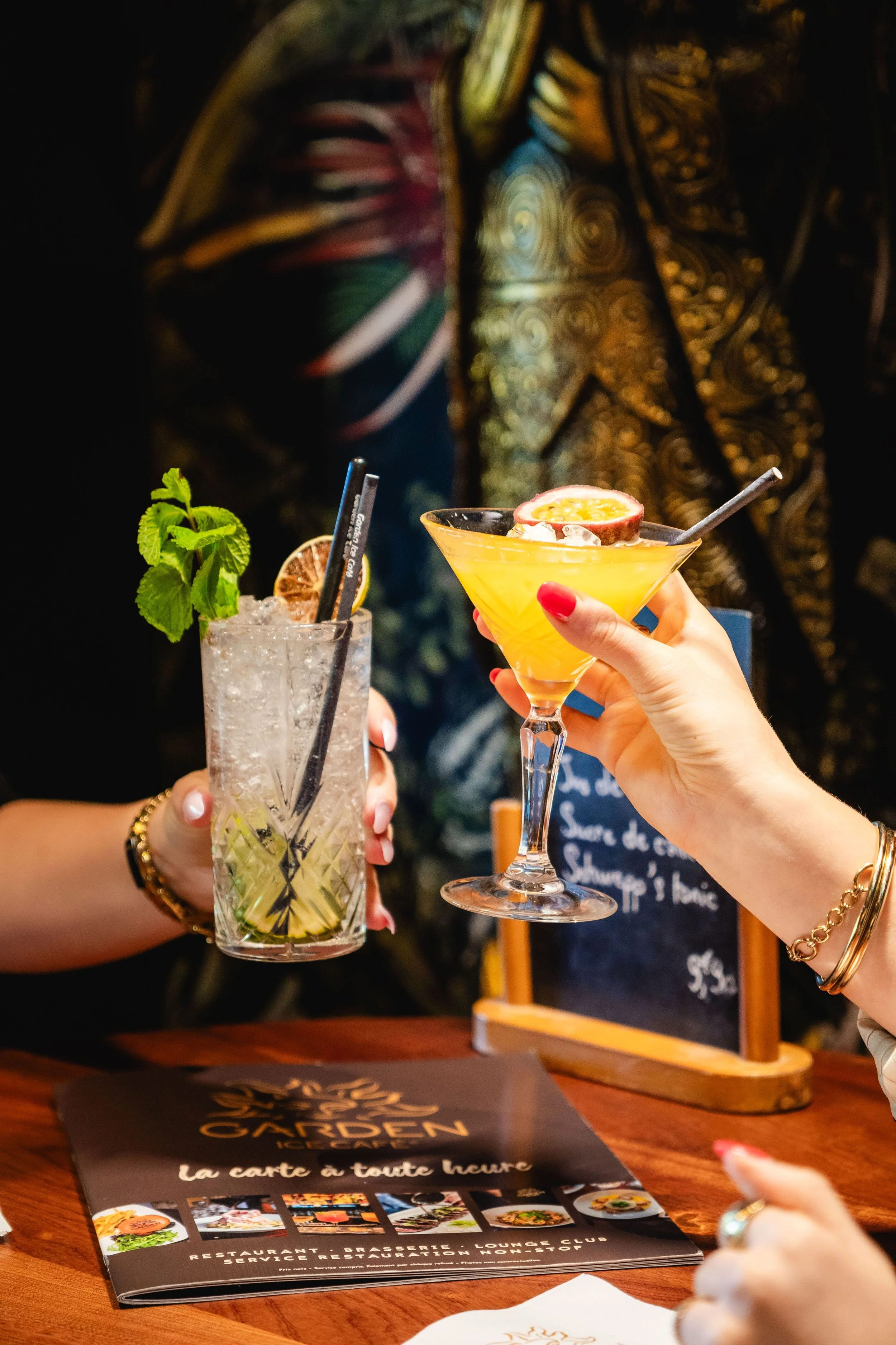 Deux personnes tenant des cocktails au bar, un cocktail avec une tranche de citron vert, un autre avec une tranche de passion, un menu sur la table, au Garden Ice Café