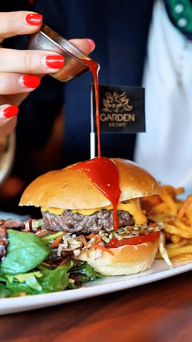Une personne verse de la sauce piquante sur un burger, accompagné de frites, sur une assiette au Garden Ice Café