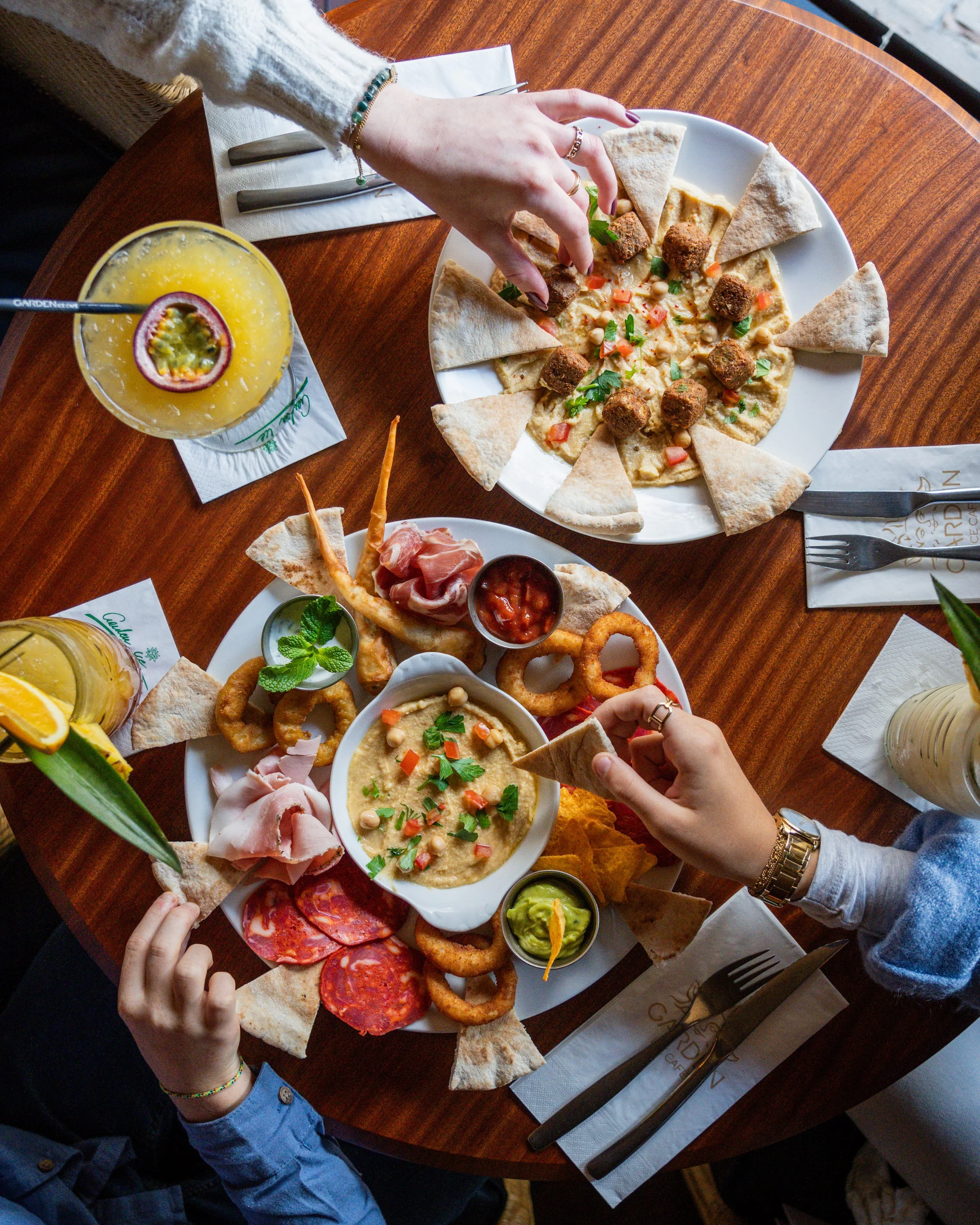 Un repas de type tapas avec différentes assiettes de nourriture, y compris du houmous, du guacamole, des chips, des légumes, de la charcuterie, des falafels, et des boissons, sur une table en bois au Garden Ice Café