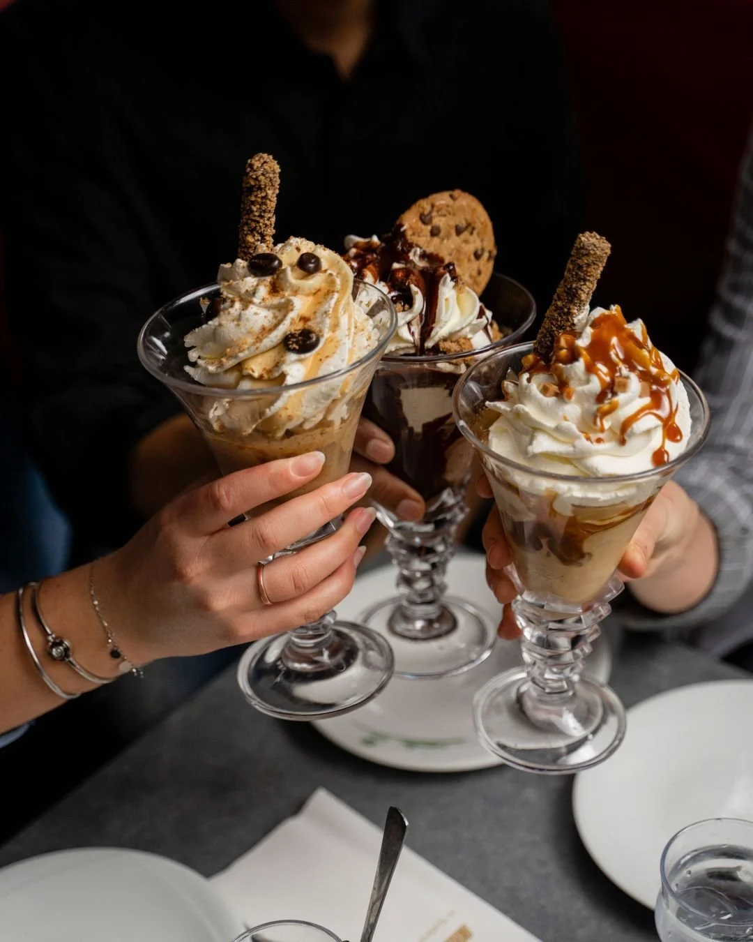 Trois mains tiennent des coupes de crème glacée avec des toppings comme des biscuits, du caramel, du chocolat, et de la crème fouettée au Garden Ice Café