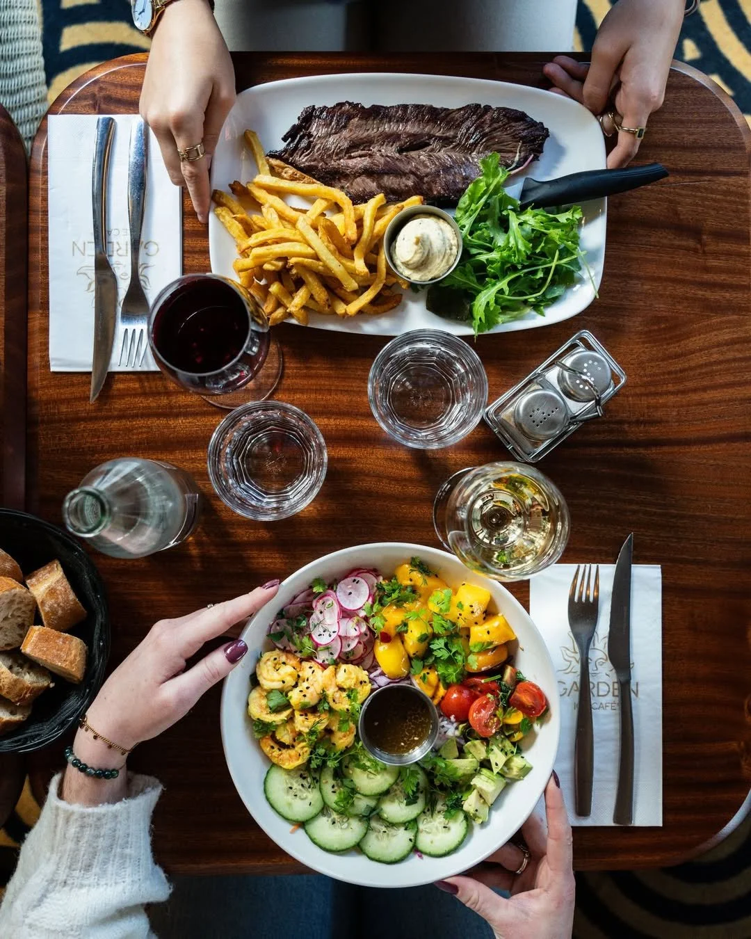 Repas avec deux plats de salade, une assiette de viande avec des frites, verres d'eau, vin blanc et rouge, ainsi qu'un panier de pain sur une table en bois au Garden Ice Café
