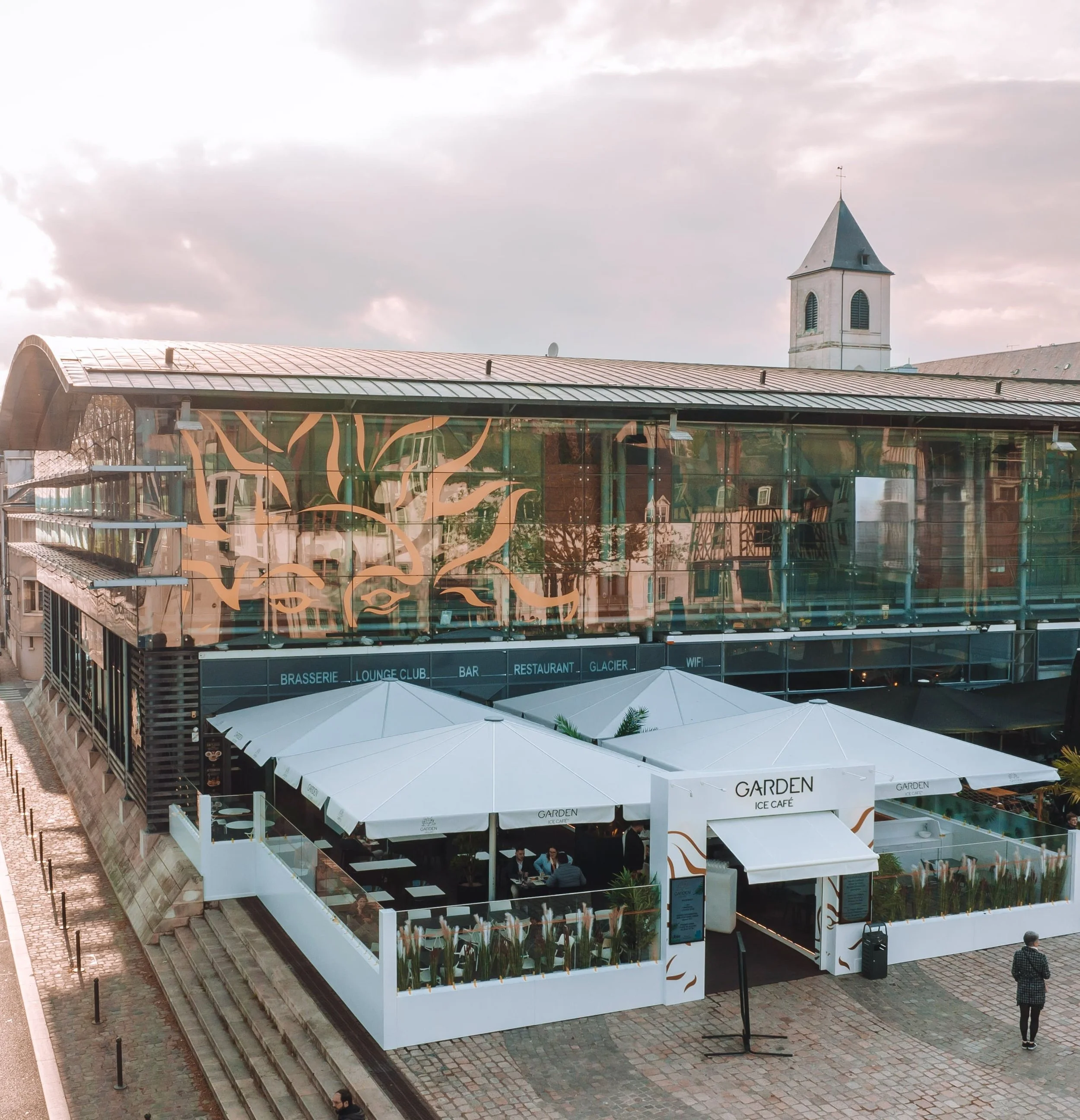 Un bâtiment moderne avec une façade en verre, une terrasse avec des parasols blancs, un panneau indiquant 'GARDEN ICE CAFE'