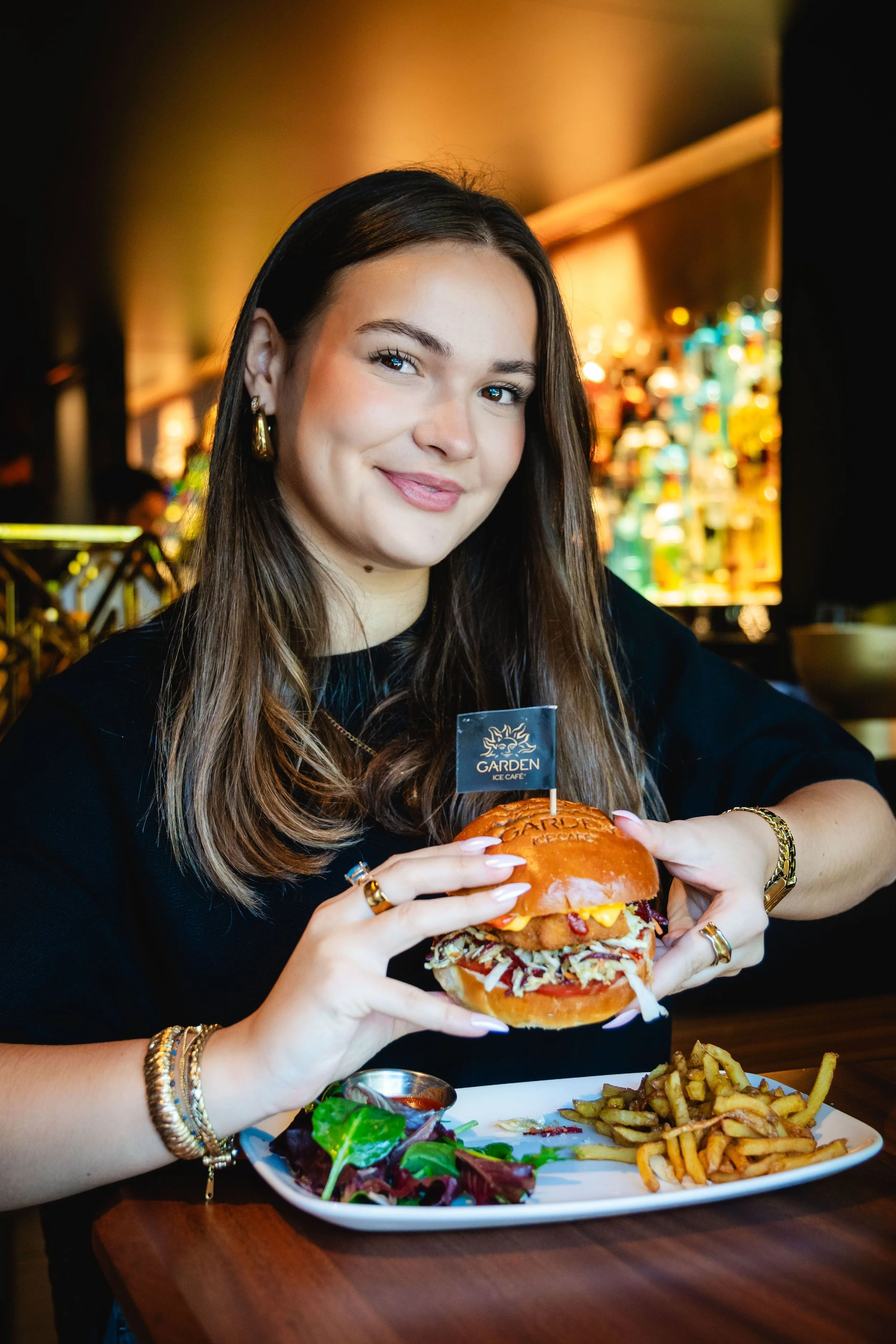 Jeune femme souriante tenant un hamburger au Garden Ice, avec salade et frites sur assiette devant elle.