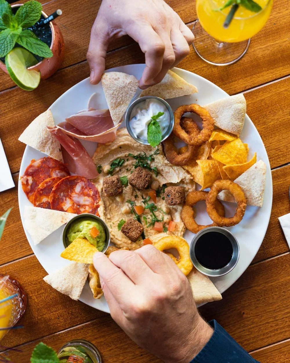 Assiette de nourriture variée comprenant des tranches de jambon, des olives, du houmous avec des morceaux de légumes, des rondelles d'oignons frits, du guacamole, du pain pita coupé en triangles, des chips, des sauces, au garden Ice