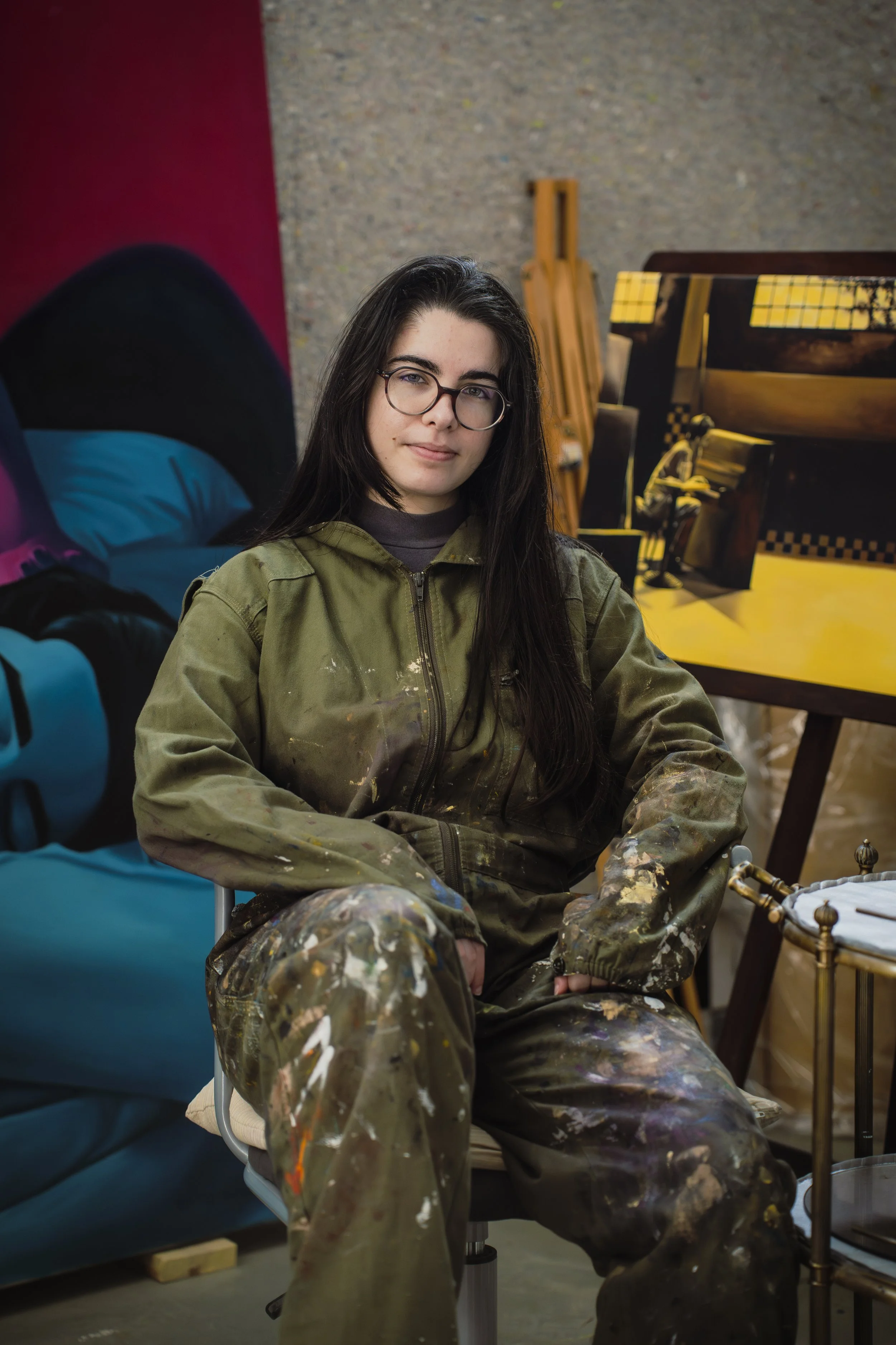 Jovem artista com cabelo comprido e óculos, vestindo roupa de pintar cheia de manchas de tinta, sentada em cadeira na frente de um atelier de arte com pintura ao fundo.