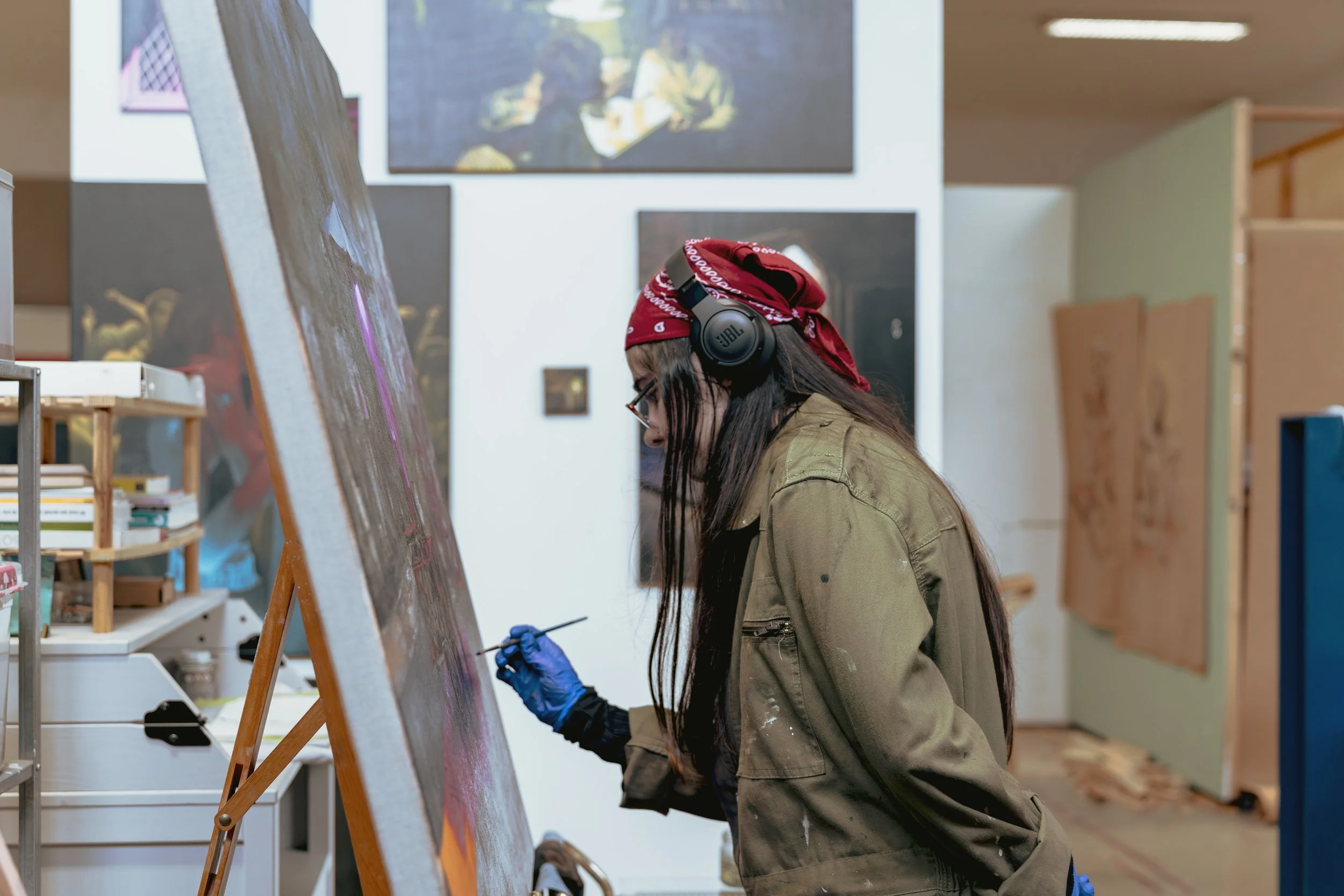 Mulher pintando em um ateliê de arte, usando óculos, bandana vermelha, jaqueta verde, luvas azuis e fones de ouvido enquanto pinta uma tela grande.