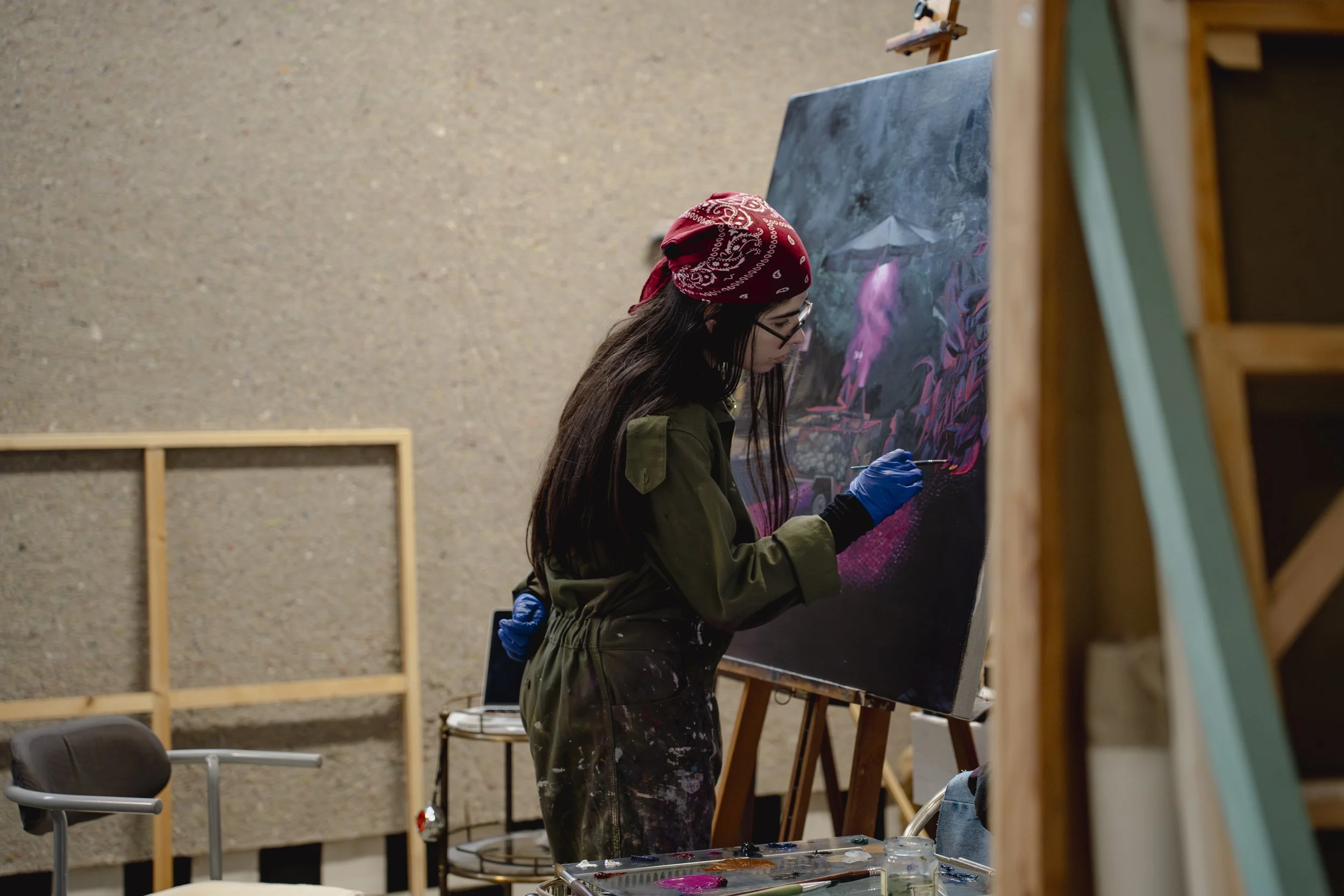 Mulher artista pintando uma tela em um estúdio de arte, usando luvas azuis e usando um lenço vermelho na cabeça.