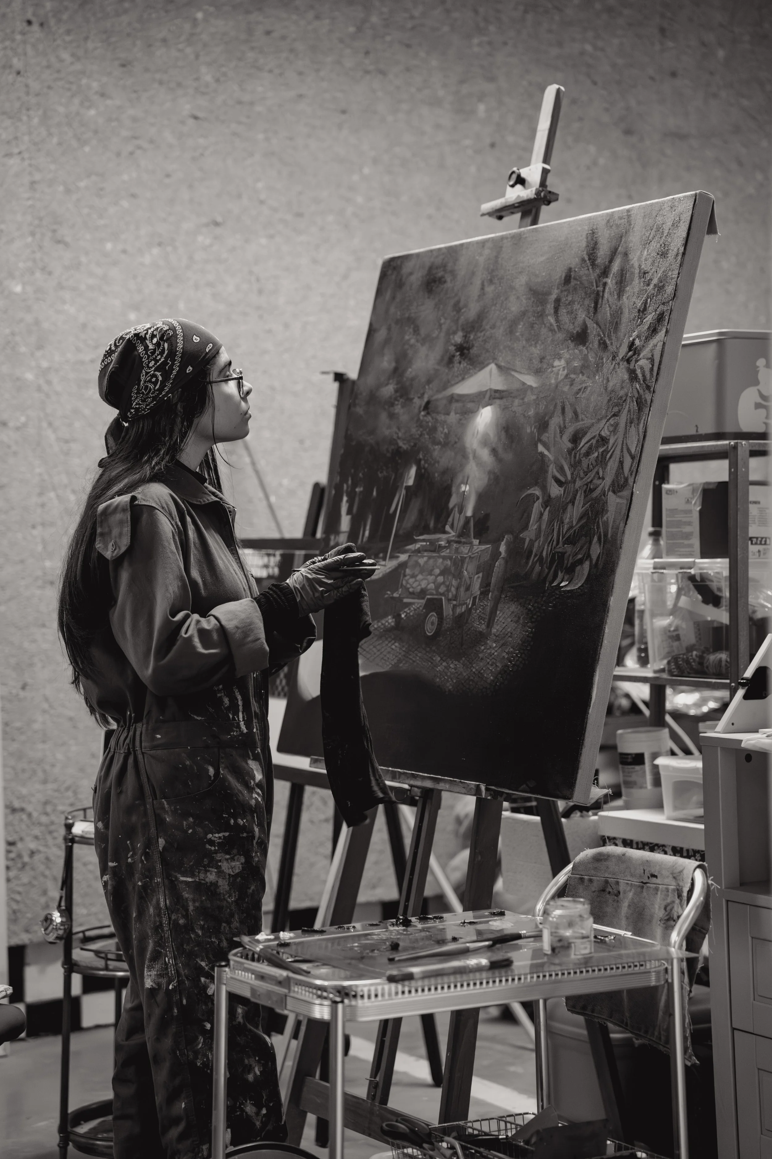 Beatriz Narciso pintando um quadro no seu estúdio, usando um avental manchado de tinta, com uma mesa de trabalho com pincéis e tinta ao seu lado.