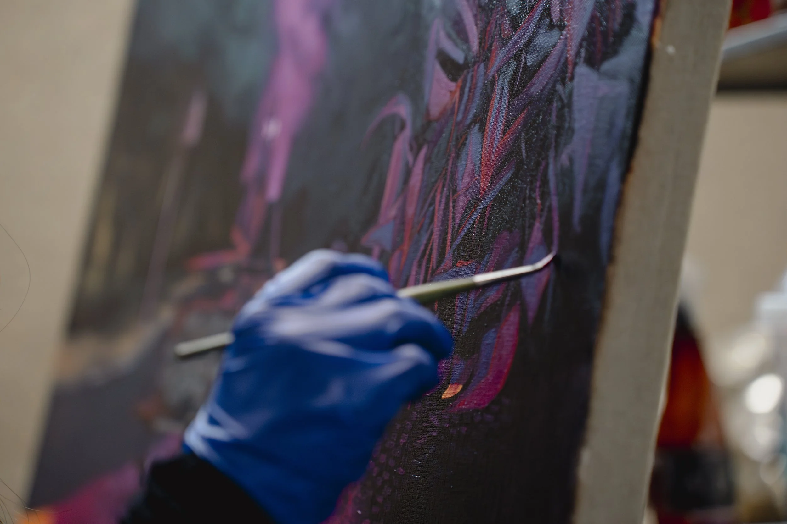 Pessoa usando luva azul pintando um quadro com tinta em um cavalete, com destaque para detalhes de cores vibrantes na pintura.