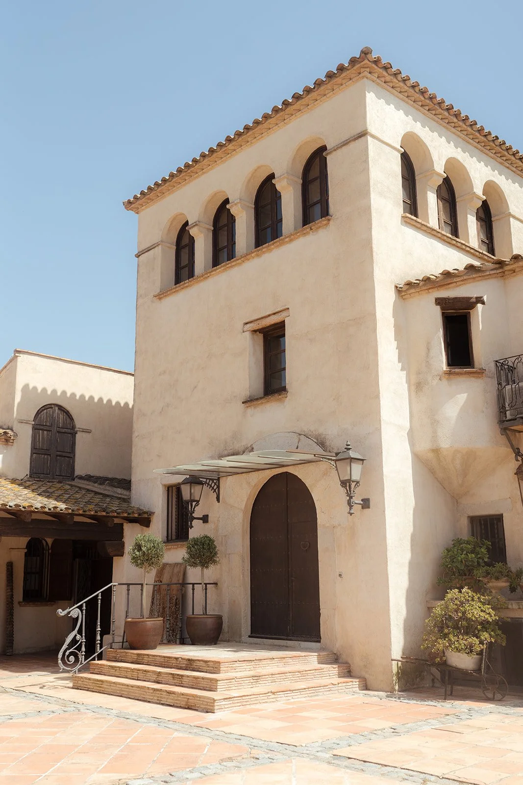 A Mediterranean-style building with beige stucco walls, arched windows, and a tiled roof. There is a large wooden door at the entrance, flanked by two outdoor lamps and potted plants on steps leading up to it. The courtyard has terracotta tiles and additional potted greenery.