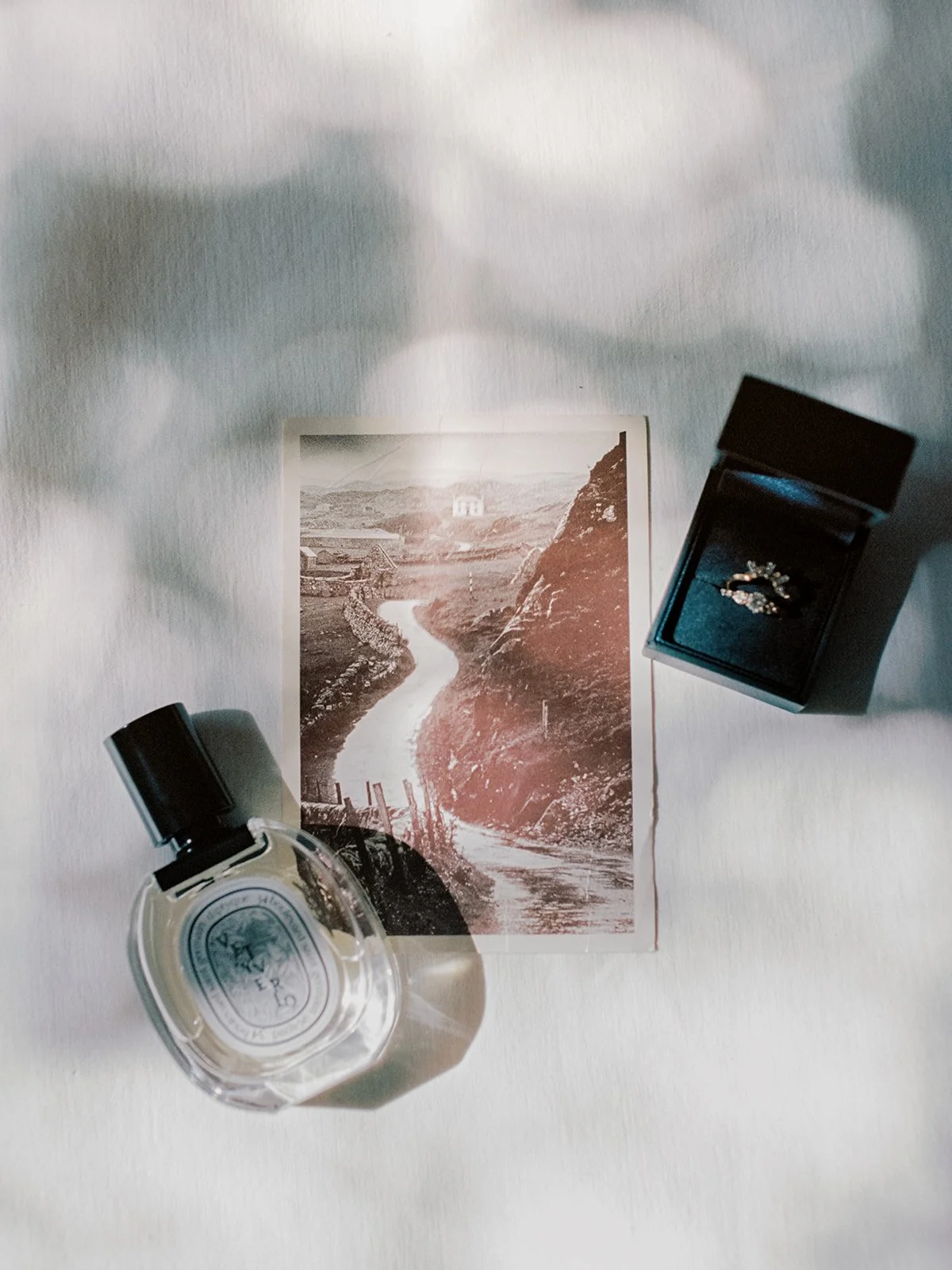 A black-and-white photograph of a river winding through a valley, with hills and a small building in the background. A perfume bottle and an open jewelry box with a ring are placed on the photograph.