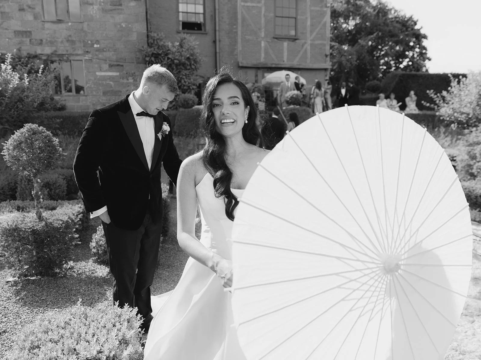A bride in a white wedding dress and a groom in a black tuxedo are outdoors, with the bride smiling at the camera and a large white parasol partially covering her. The groom is looking down, adjusting his sleeve. There are people and a building in the background.