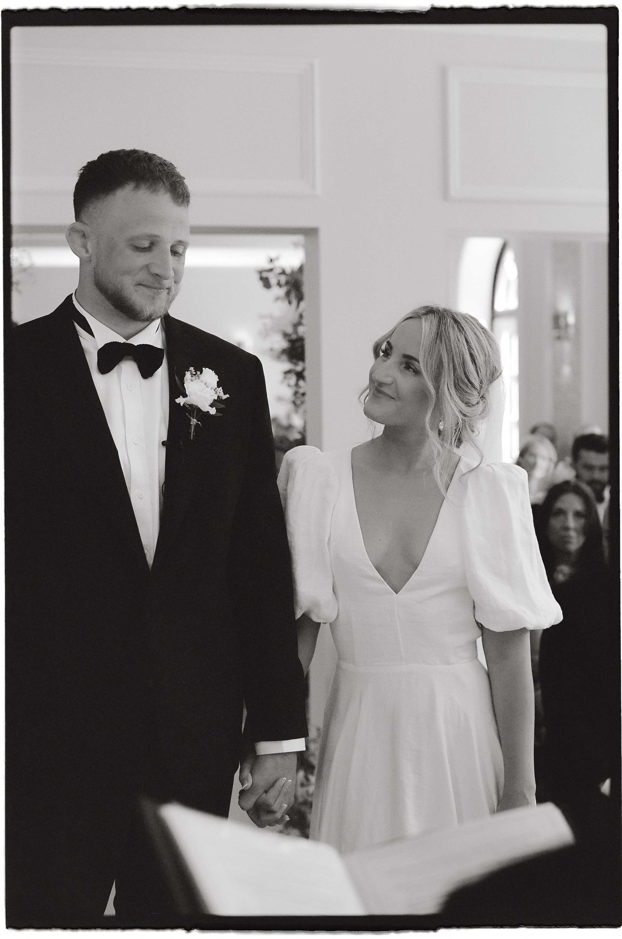 A black and white photo of a wedding ceremony showing a bride and groom holding hands. The groom wears a tuxedo with a bow tie and boutonnière, and the bride in a white dress with puffed sleeves looks at him lovingly. Guests are visible in the background.