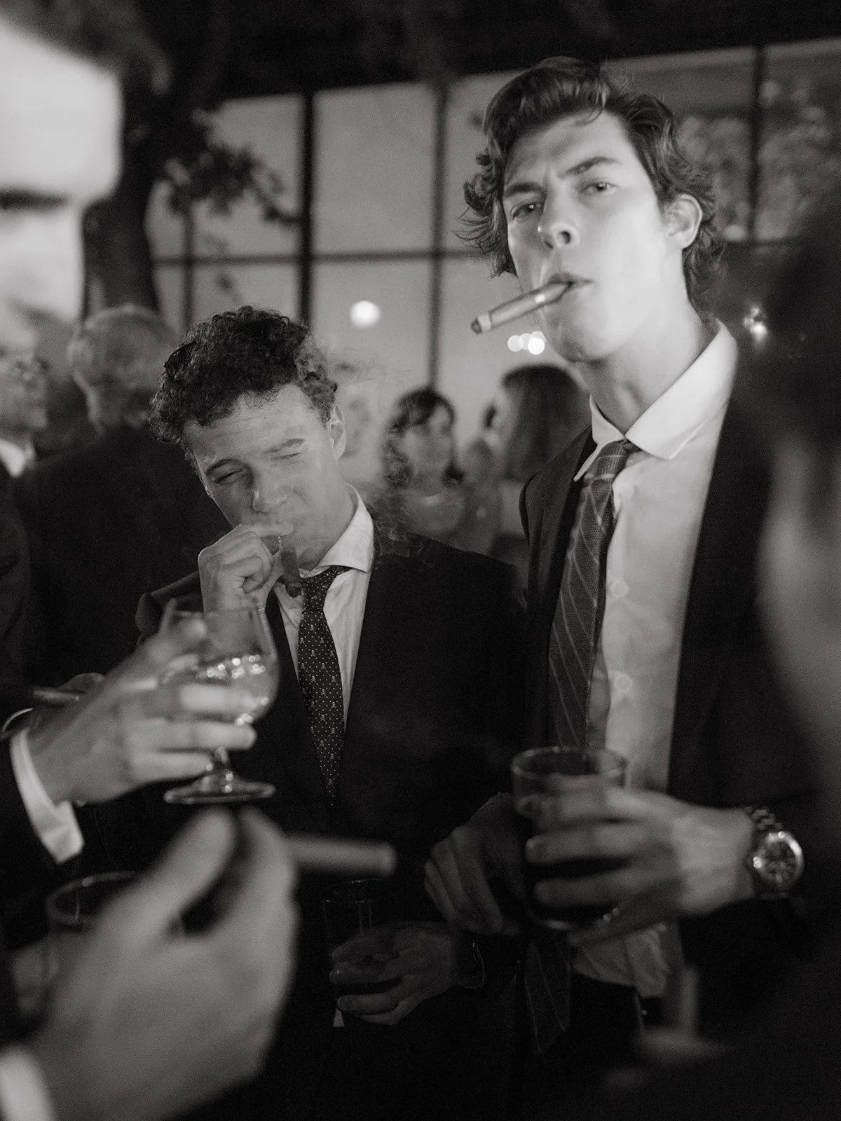 Two men in formal suits at a social gathering, one smoking a cigarette and holding a drink, the other with a cigarette, surrounded by other people in a dimly lit venue L'Orangerie de Clos Barenys, Cambrils, Spain