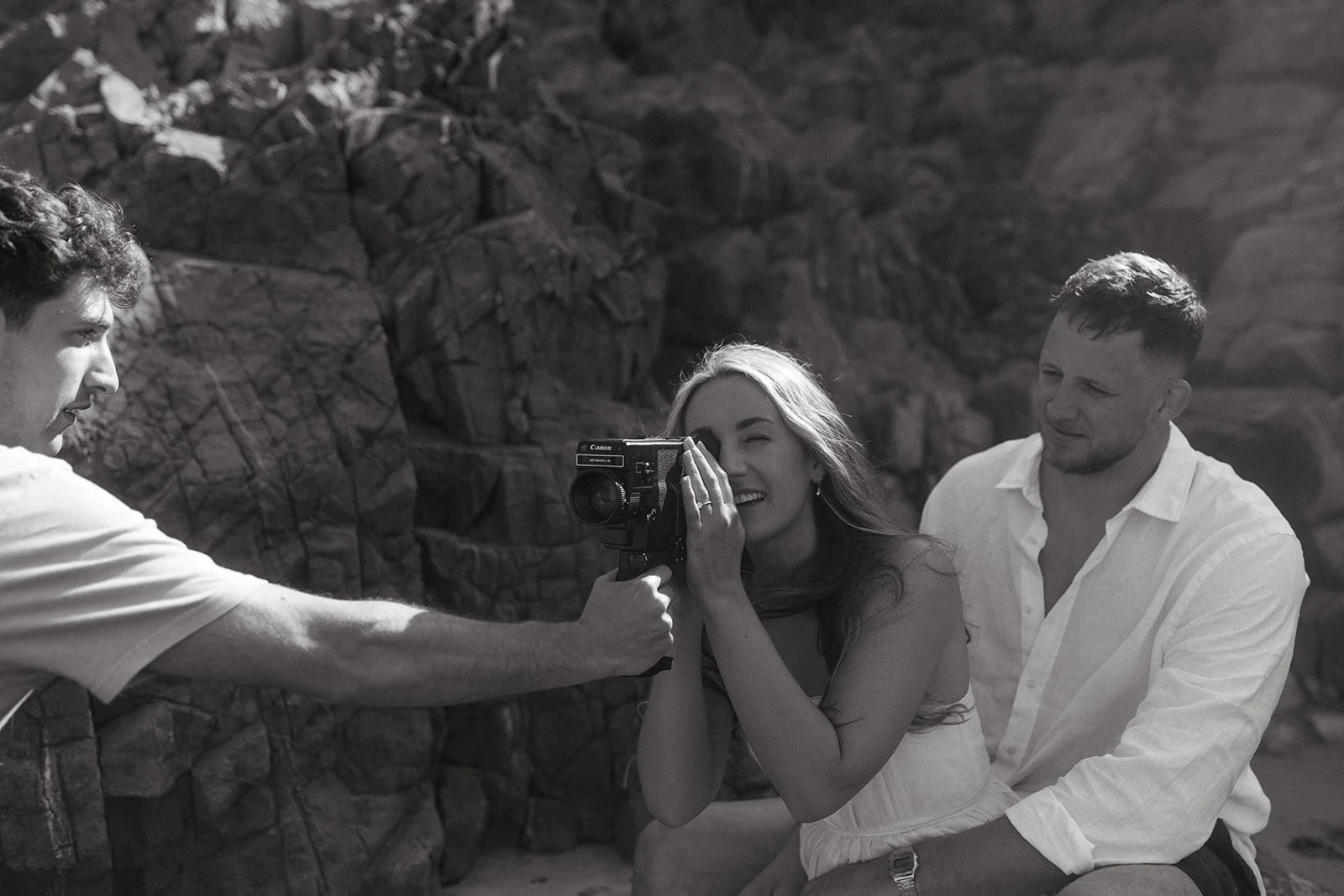 A woman with long hair smiling and looking through a camera, while a man holds the camera steady and another man sits beside her with his arms around her waist, all outdoors against a rocky background.