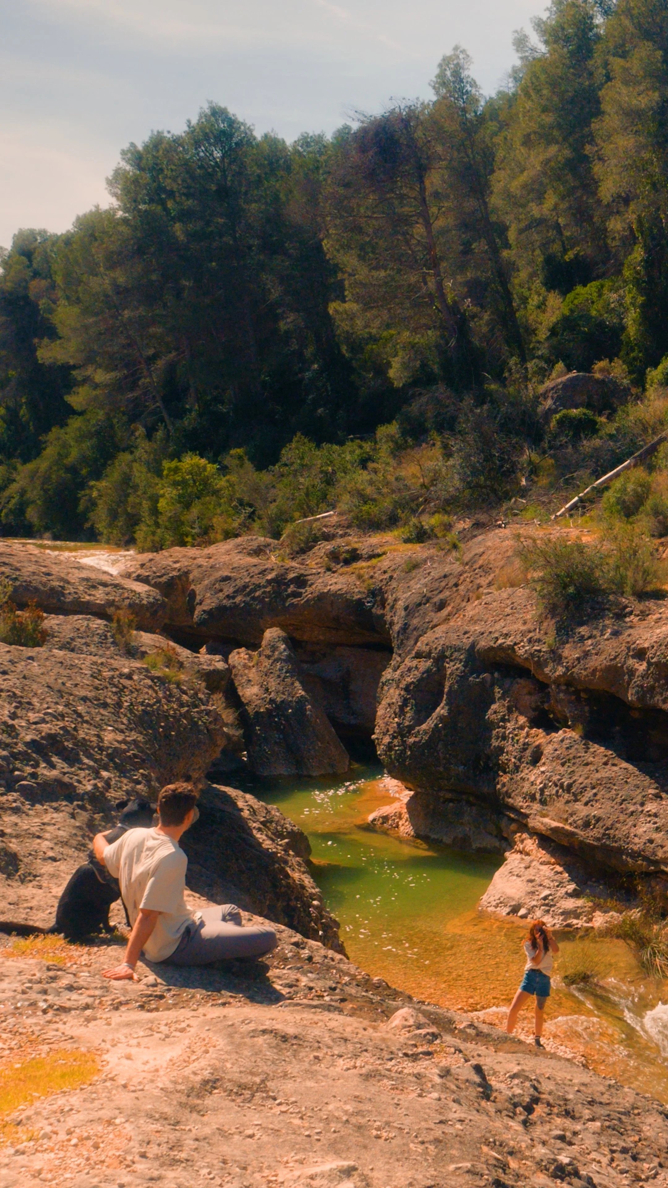 an image of a couple exploring nature in the catalonian mountains, one person is sat with dog and the other person is taking a photograph of the river