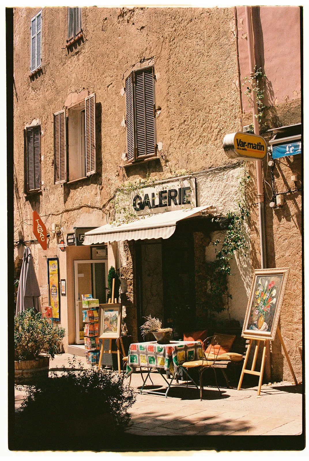 Outdoor scene of a European street with an art gallery, tables, and paintings displayed outside, under sunlight.