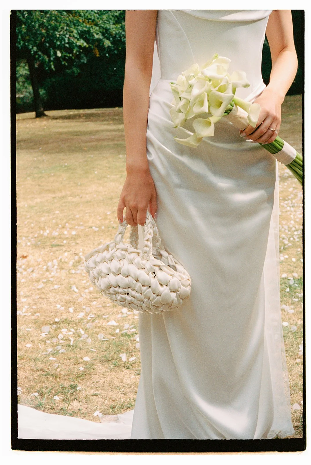 A woman in a white satin wedding dress holding a bouquet of white calla lilies and a woven white purse, standing outdoors on a grassy area with trees in the background.