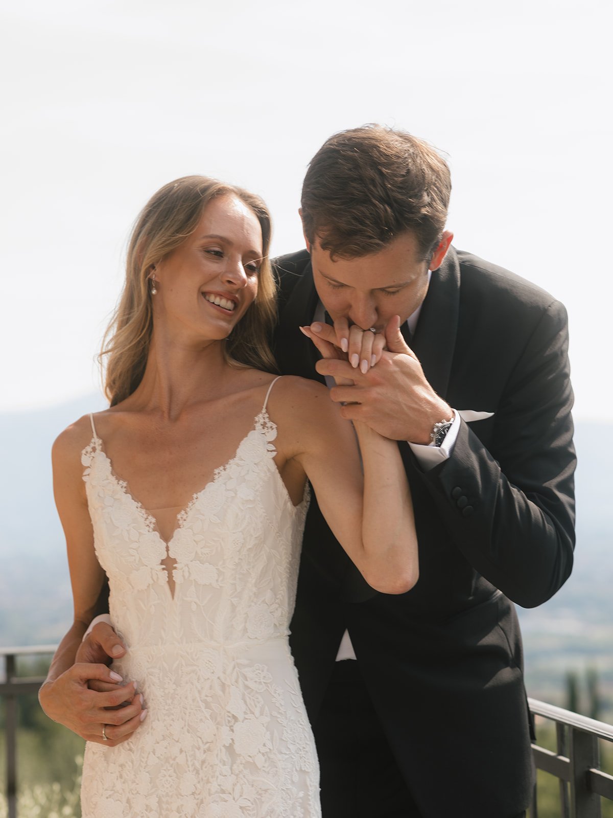 A smiling bride in a white lace wedding dress being kissed on the hand by a groom in a black tuxedo outside with a scenic view of mountains and sky.