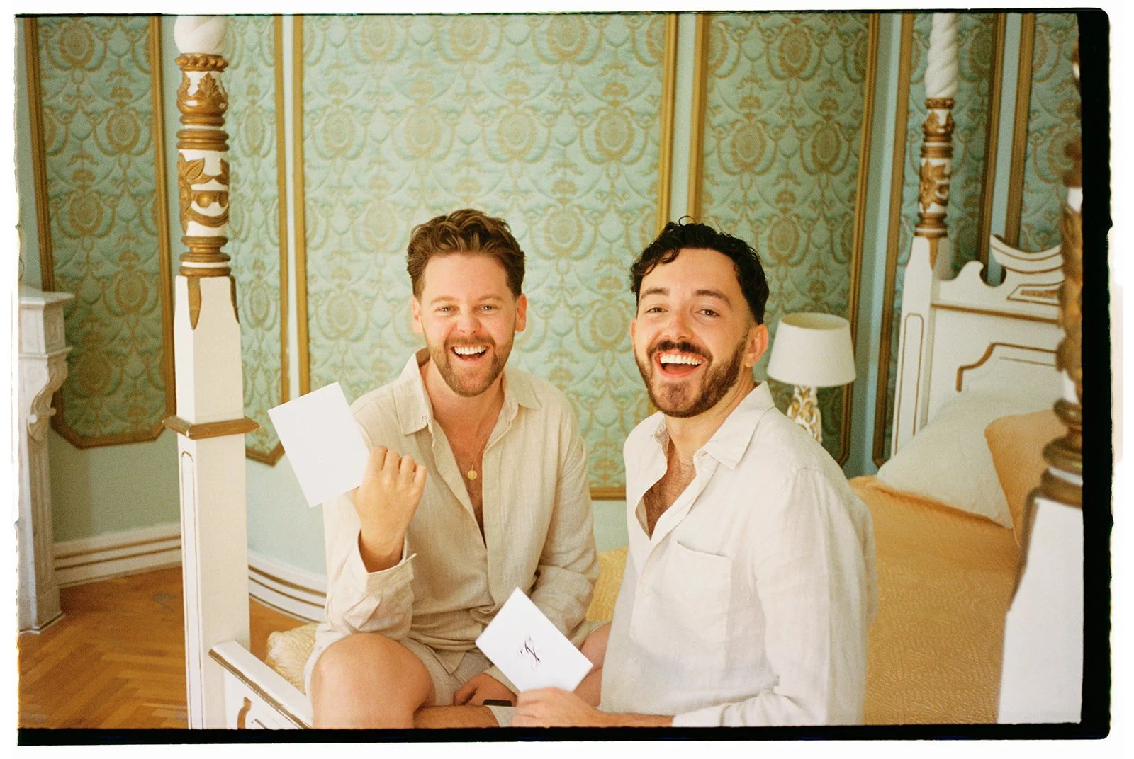 Two smiling men sitting on a bed in a luxurious, ornately decorated bedroom, holding white sheets of paper.