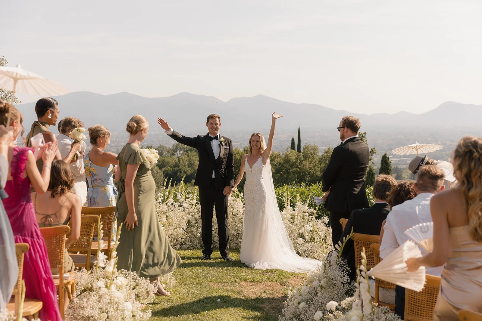 A newlywed couple holding hands and smiling at their wedding ceremony outdoors, with guests standing and seated on either side, overlooking a scenic valley with mountains in the background.