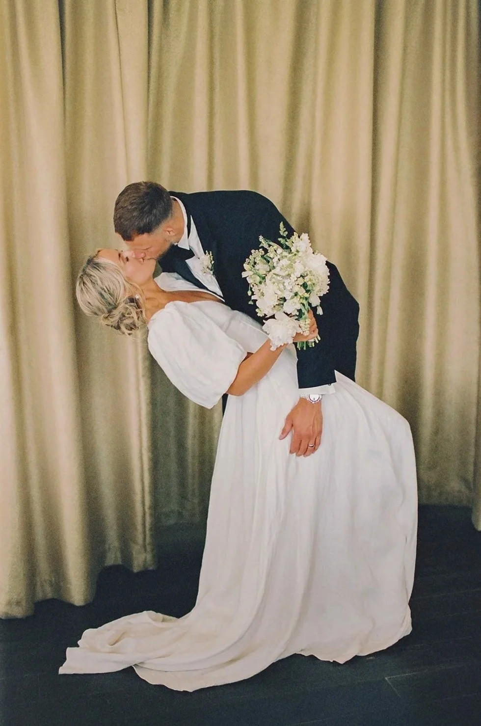 A bride and groom share a kiss, with the groom dipping the bride while holding a bouquet of white flowers. The bride wears a white dress and the groom a tuxedo. They are standing in front of gold-colored curtains.
