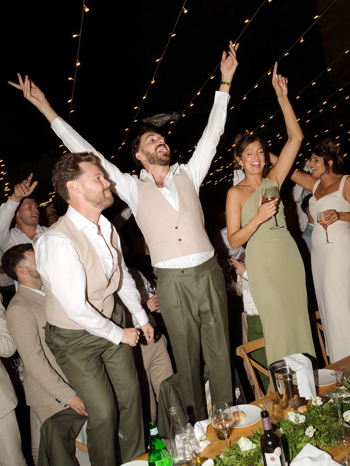 People celebrating at an event, with a man raising his arms in joy, surrounded by other smiling guests holding wine glasses, under string lights at night.