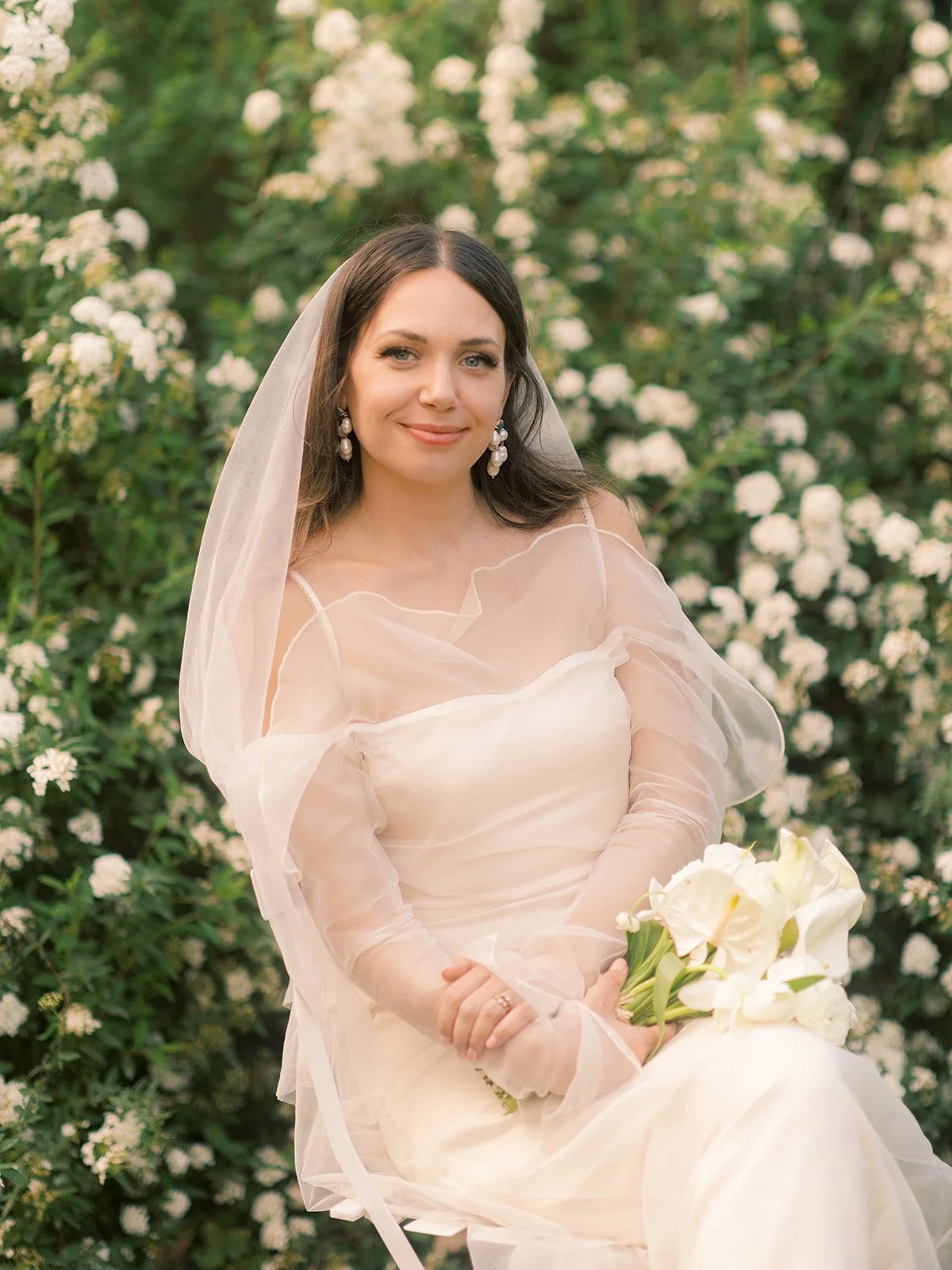A woman in a wedding dress sitting outdoors in front of white flowering bushes, holding a bouquet of white flowers, and smiling at the camera in Nîmes, South Of France.