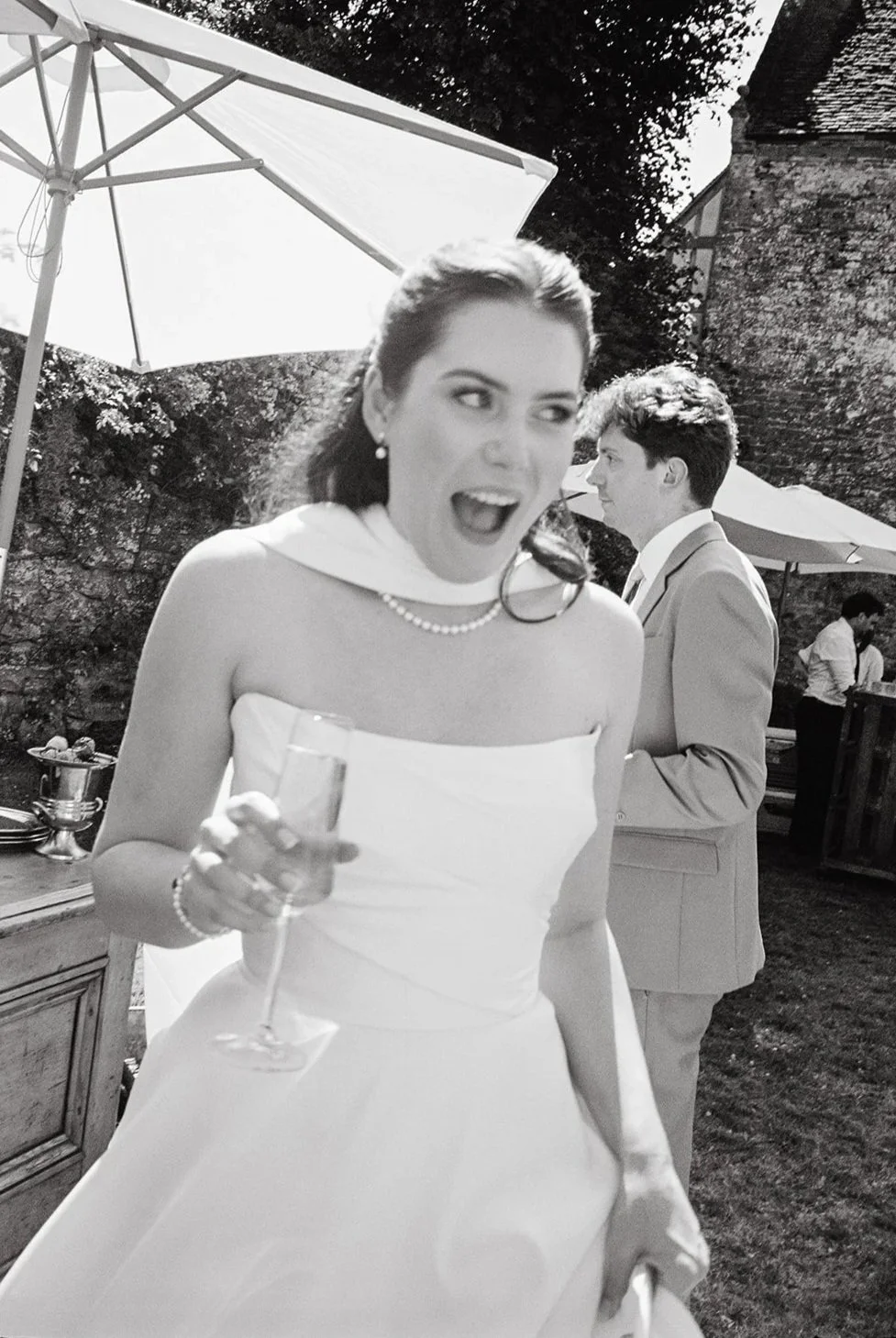 A woman in a white dress holding a champagne glass, smiling and appearing to be happy at an outdoor event, with another man in a suit in the background and umbrellas providing shade.