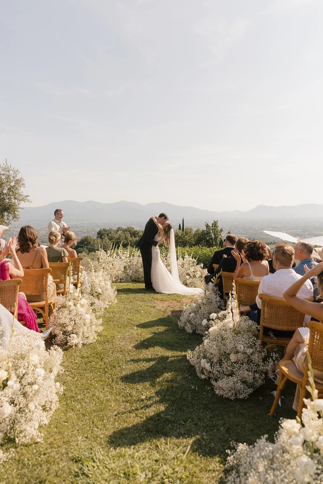 A wedding ceremony outdoors with a couple kissing, surrounded by seated guests, floral arrangements, and scenic mountains in the background.