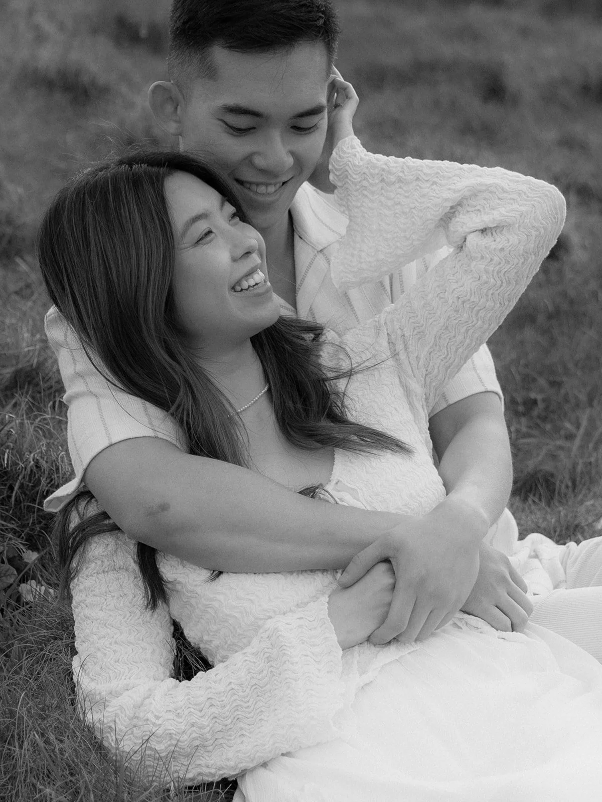 A smiling couple sitting on grass, with the woman leaning against the man, both laughing and enjoying each other's company outdoors.