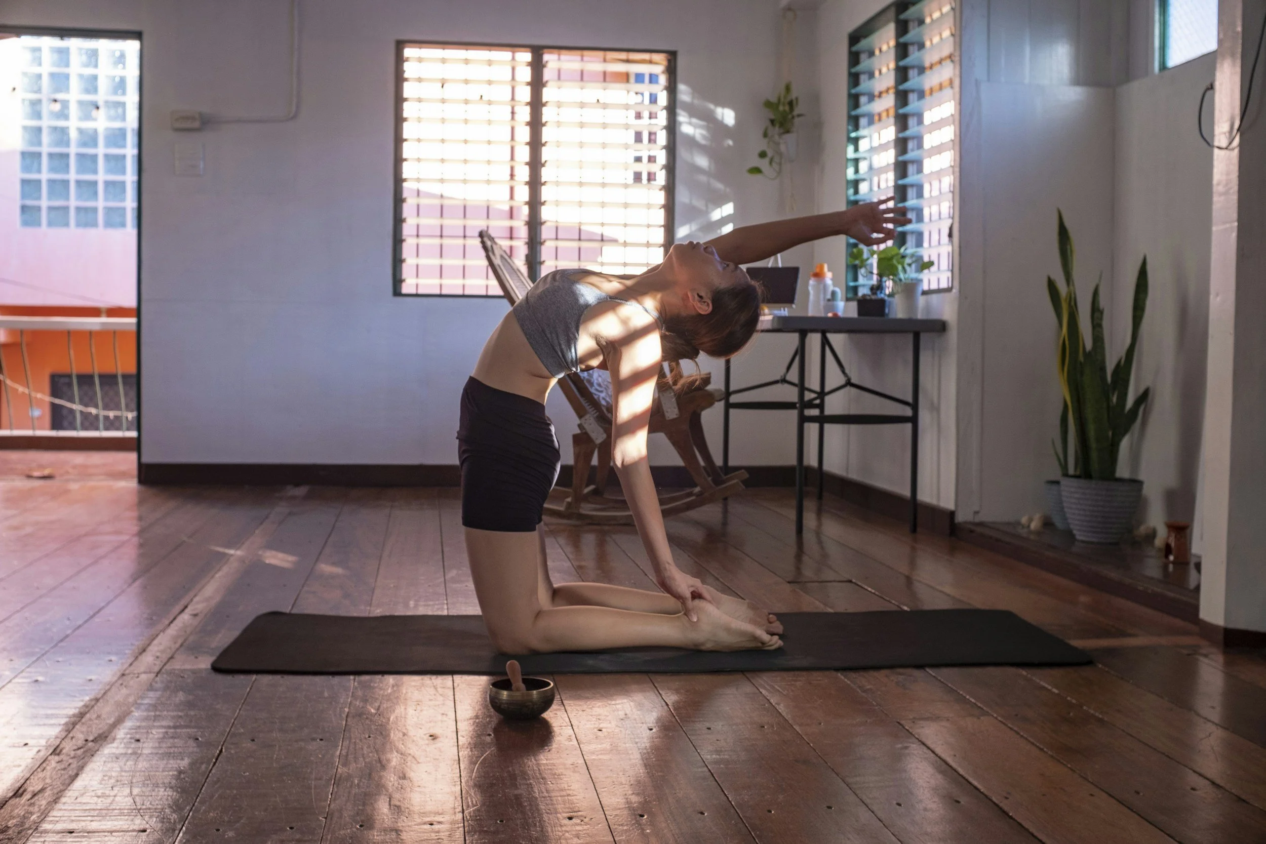A woman practicing yoga indoors, in a pose where she is on her stomach with arms and legs raised, in front of a window with blinds, in a room with wooden floors and minimal furniture.