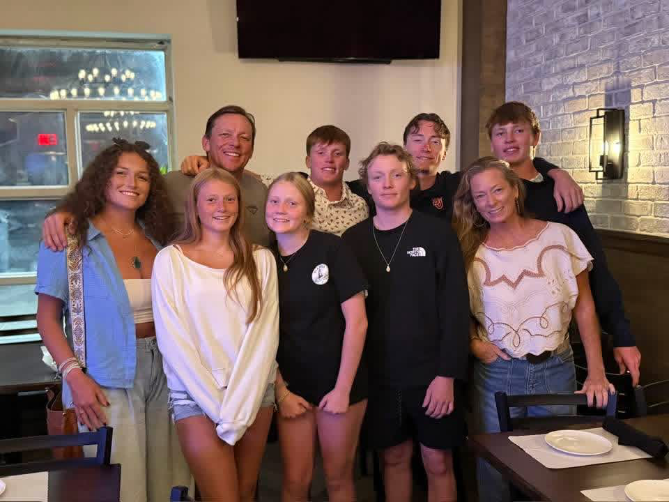 Group of nine young people and two adults, smiling and posing for a photo in a restaurant or cafe with brick walls.