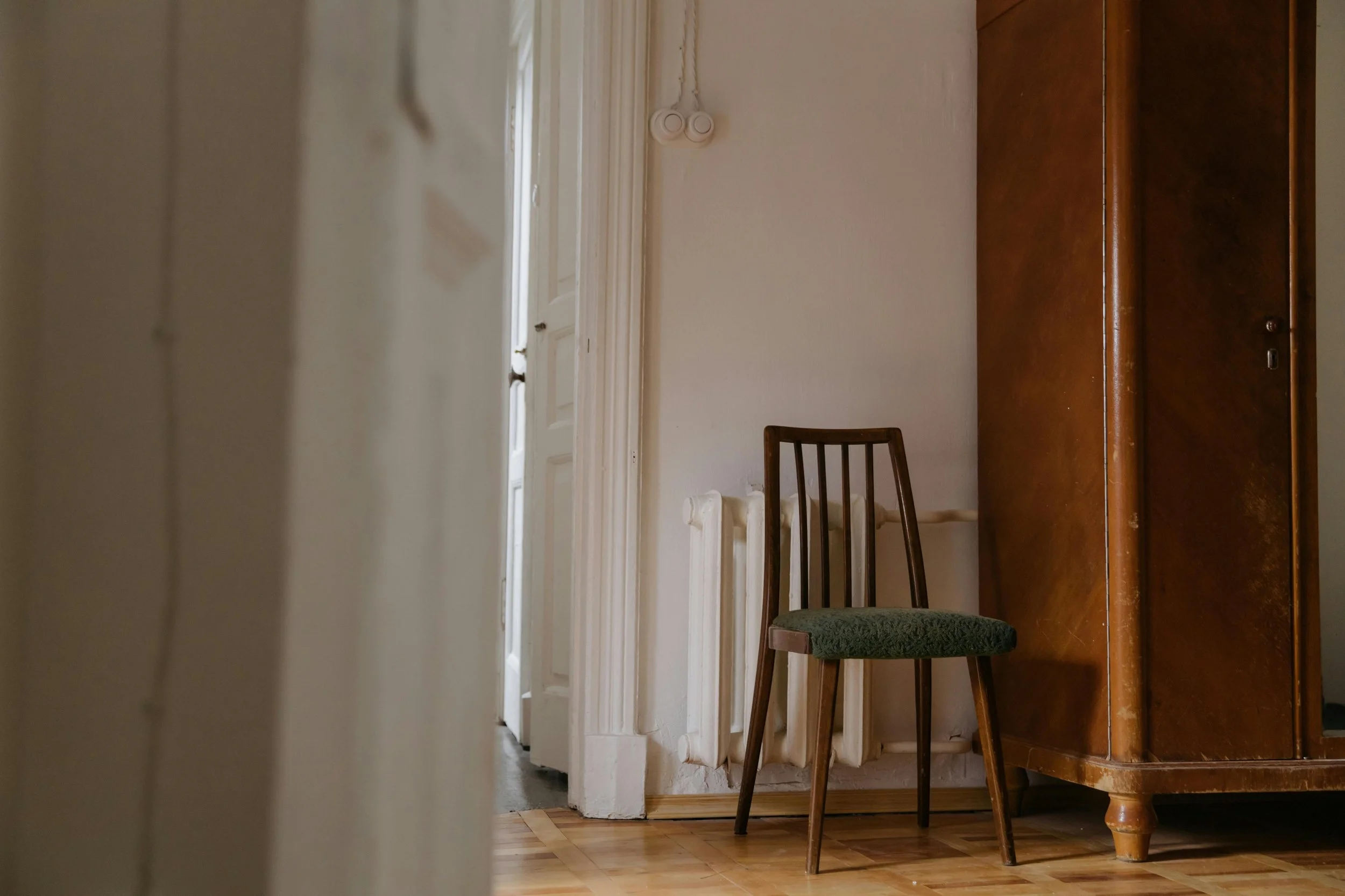 Une pièce intérieure avec une chaise en bois avec un coussin vert, une armoire en bois foncé et un radiateur blanc, vue partielle par une ouverture ou porte entrouverte.
