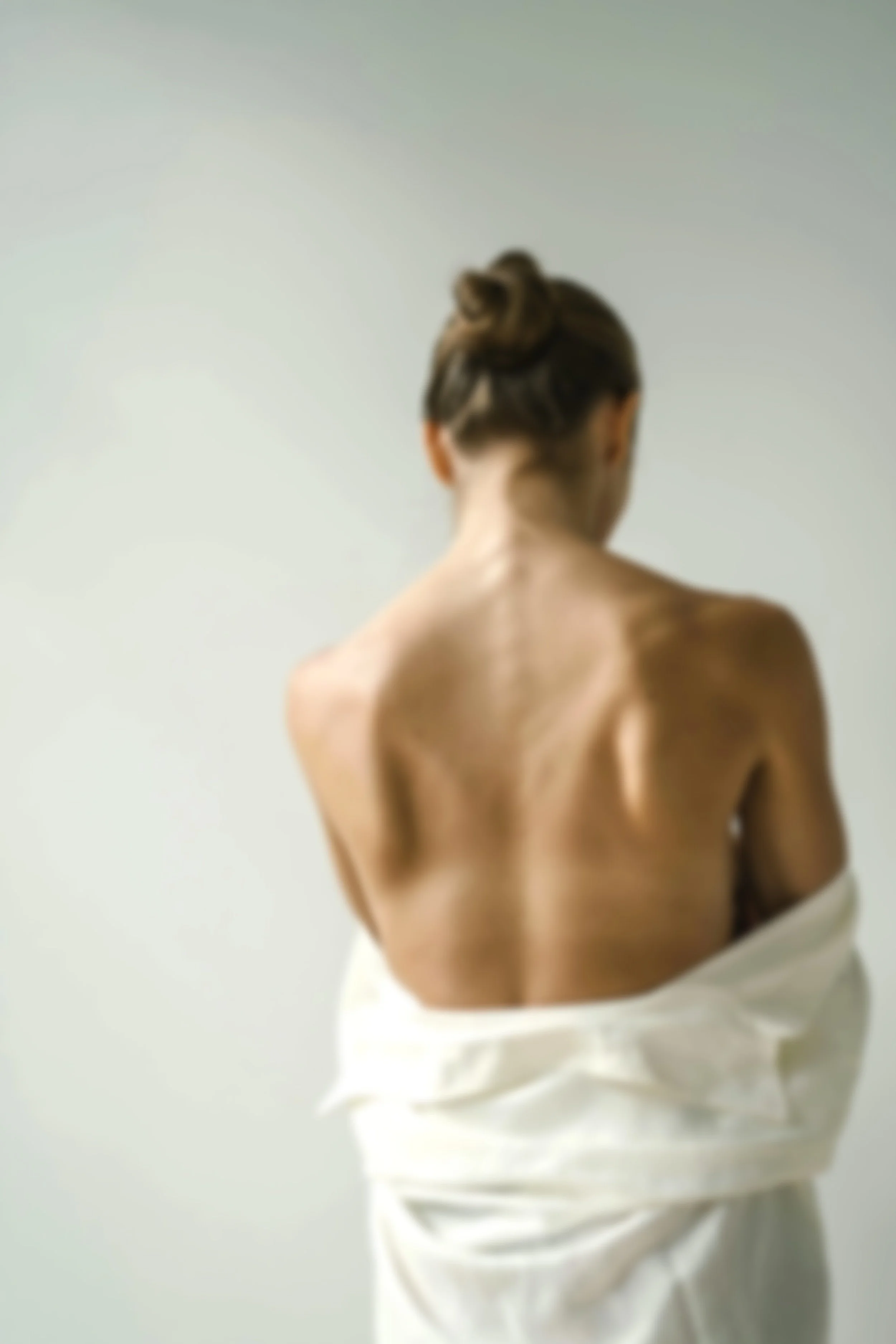 Une femme vue de dos, portant une serviette blanche autour de la taille, avec les épaules nues, fond neutre.