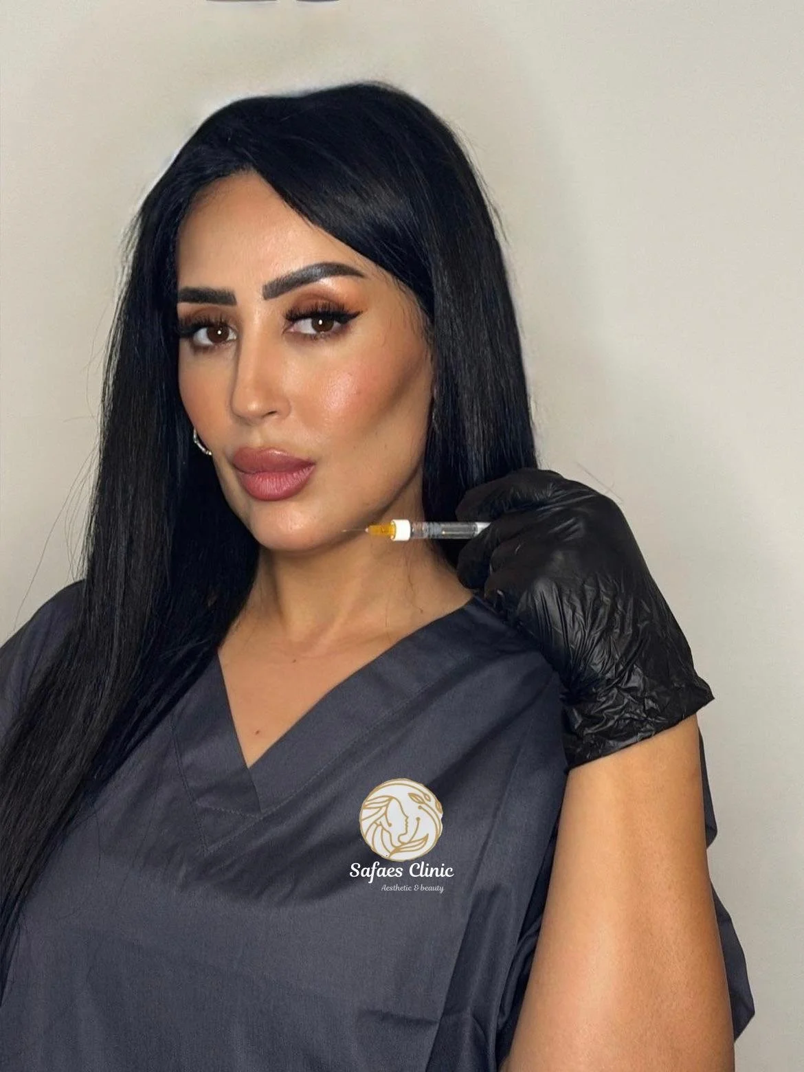 A woman with long black hair, wearing makeup, holding a syringe near her cheek, dressed in a dark clinical uniform with the logo 'Safae's Clinic Aesthetic & Beauty' on it, and wearing a black glove.