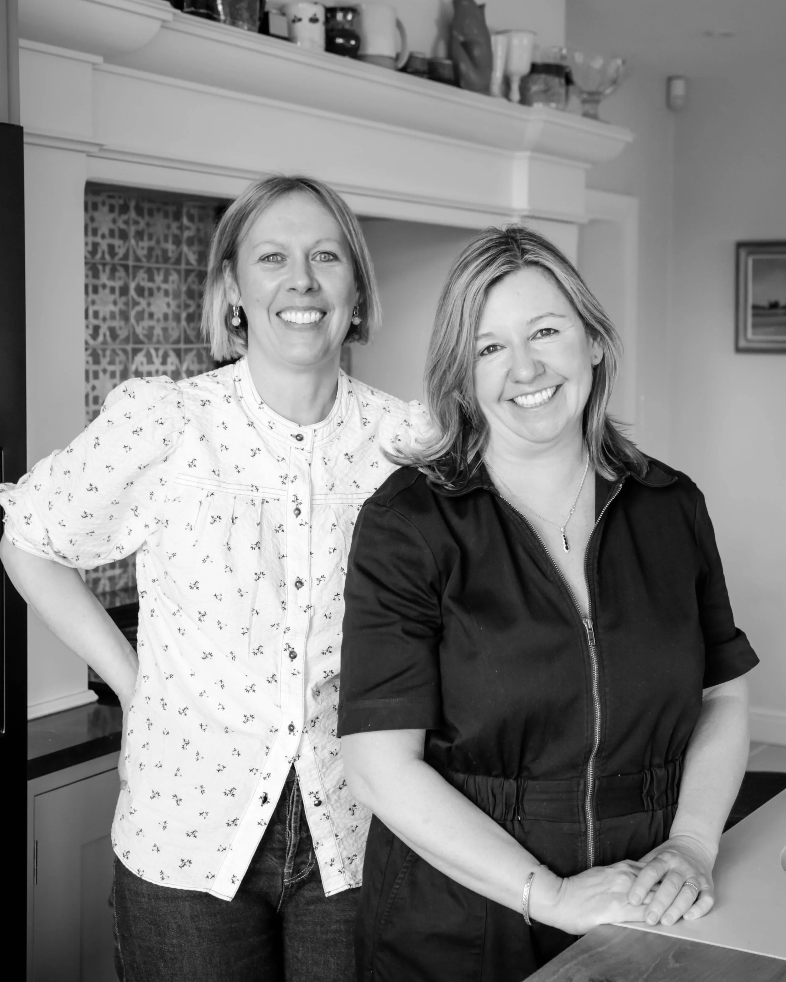 Two women smiling and posing inside a kitchen with cabinets and a decorative tile backsplash. One woman is standing and wearing a patterned blouse, the other is sitting and wearing a dark top, with her hands resting on a table or counter.