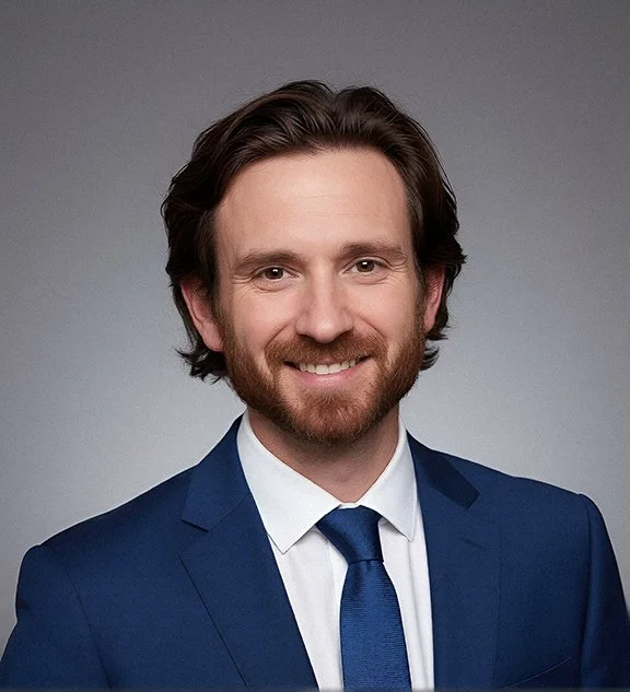 A man with brown hair and a beard, smiling, wearing a blue suit with a white shirt and blue tie, against a gray background.
