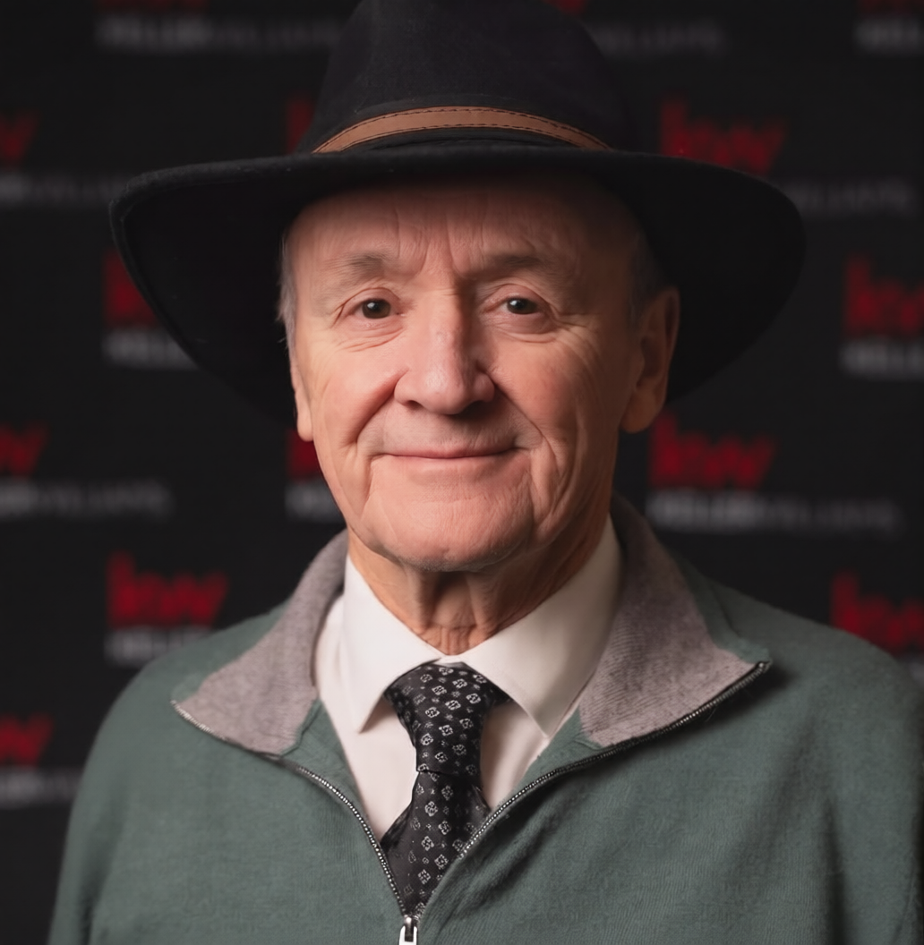 An elderly man wearing a black hat, gray jacket with a zipper, white shirt, and black patterned tie, smiling at the camera with a blurred black and red background.